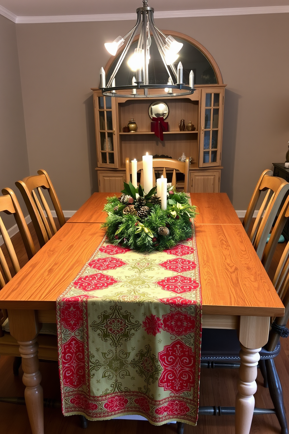 A vibrant festive table runner drapes elegantly across a polished wooden dining table, adorned with intricate patterns of red and green. Surrounding the table are mismatched chairs, each decorated with small wreaths tied with gold ribbons, enhancing the cozy holiday atmosphere. The dining space is illuminated by soft, warm lighting from a modern chandelier above, casting a welcoming glow. A centerpiece of pinecones, candles, and seasonal foliage complements the festive table runner, creating a perfect setting for holiday gatherings.