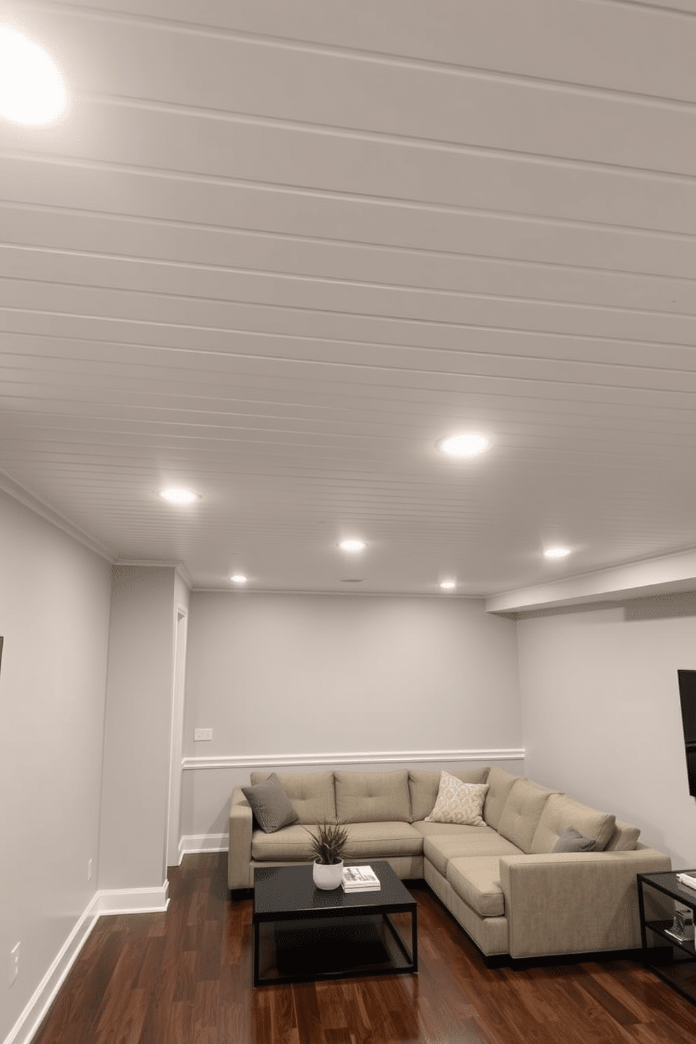 A modern basement featuring vinyl ceiling planks for durability, providing a sleek and polished look. The ceiling is complemented by recessed lighting that highlights the clean lines and enhances the spacious feel of the room. The walls are painted in a soft gray, creating a cozy atmosphere, while the flooring consists of dark hardwood that adds warmth to the space. Stylish furniture pieces, including a plush sectional sofa and a minimalist coffee table, invite relaxation and social gatherings.