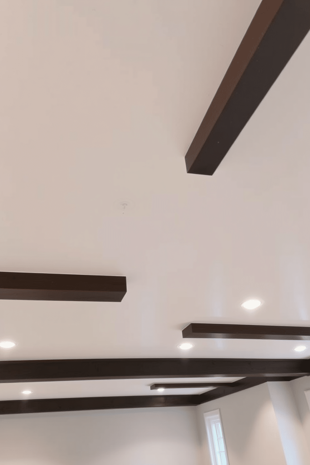 A modern basement ceiling featuring recessed lighting that creates a sleek and polished finish. The ceiling is finished in a matte white, seamlessly integrating with the minimalist design of the space. The recessed lights are strategically placed to highlight the open areas and provide ample illumination without detracting from the overall aesthetic. Dark wood beams add warmth and contrast, framing the ceiling and enhancing the contemporary feel of the basement.