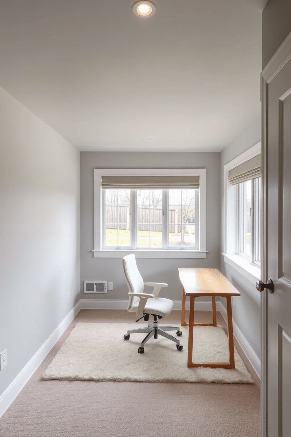 A serene basement home office features soft, muted colors that promote focus and tranquility. The walls are painted in a light dove gray, complemented by a plush area rug in a subtle cream hue. The workspace includes a sleek, minimalist desk made of natural wood, paired with a comfortable ergonomic chair in a soft fabric. Large windows allow natural light to filter in, enhancing the calming atmosphere while providing a view of the outdoor landscape.