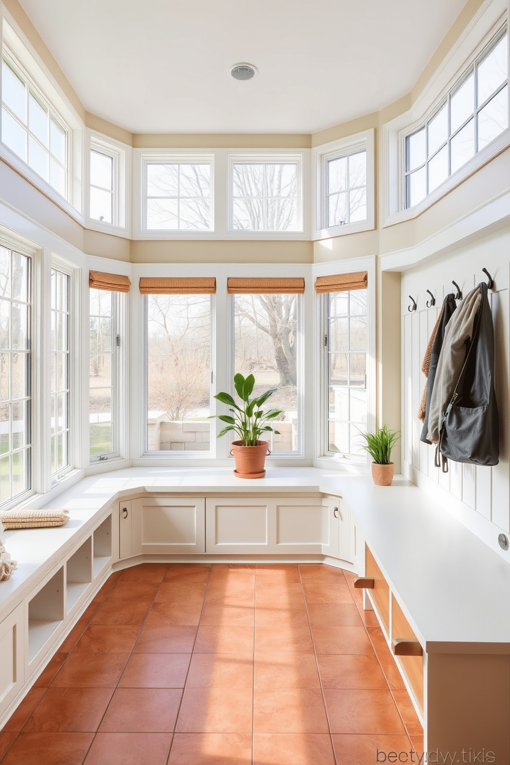 A bright and inviting mudroom design featuring large windows that flood the space with natural light. The room includes built-in storage benches with soft cushions, and hooks for coats and bags line the walls. The floor is finished with durable, weather-resistant tiles in a warm earth tone, complementing the natural light. Potted plants are placed near the windows to enhance the fresh, airy feel of the space.