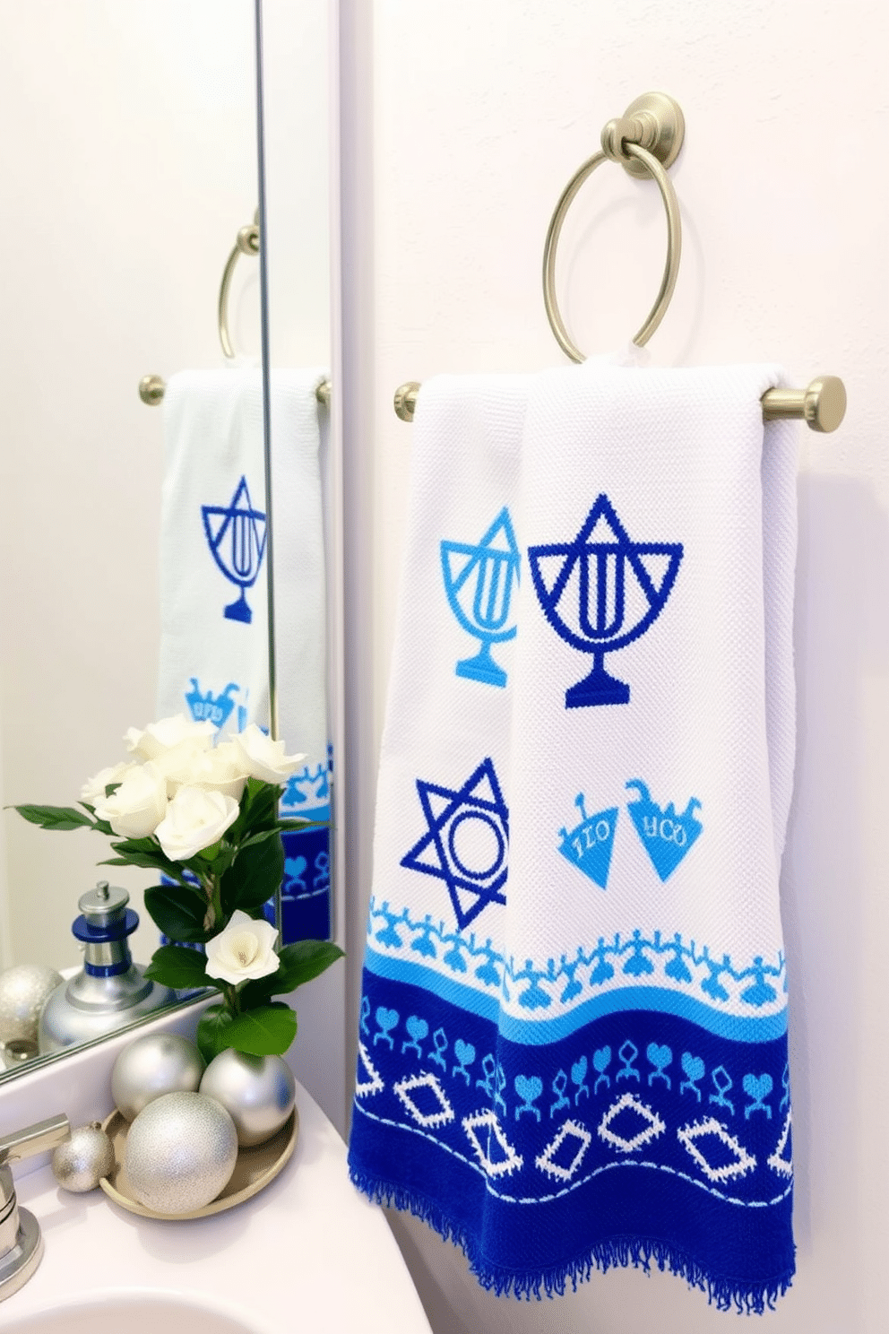 A festive bathroom setting adorned with Hanukkah-themed hand towels. The towels feature vibrant blue and white designs, showcasing traditional symbols like the menorah and dreidel, neatly hung on a decorative rack. The walls are painted a soft, wintry white, creating a bright backdrop for the holiday decor. A tasteful arrangement of silver and blue ornaments sits on the countertop, complemented by a small potted plant with white flowers.