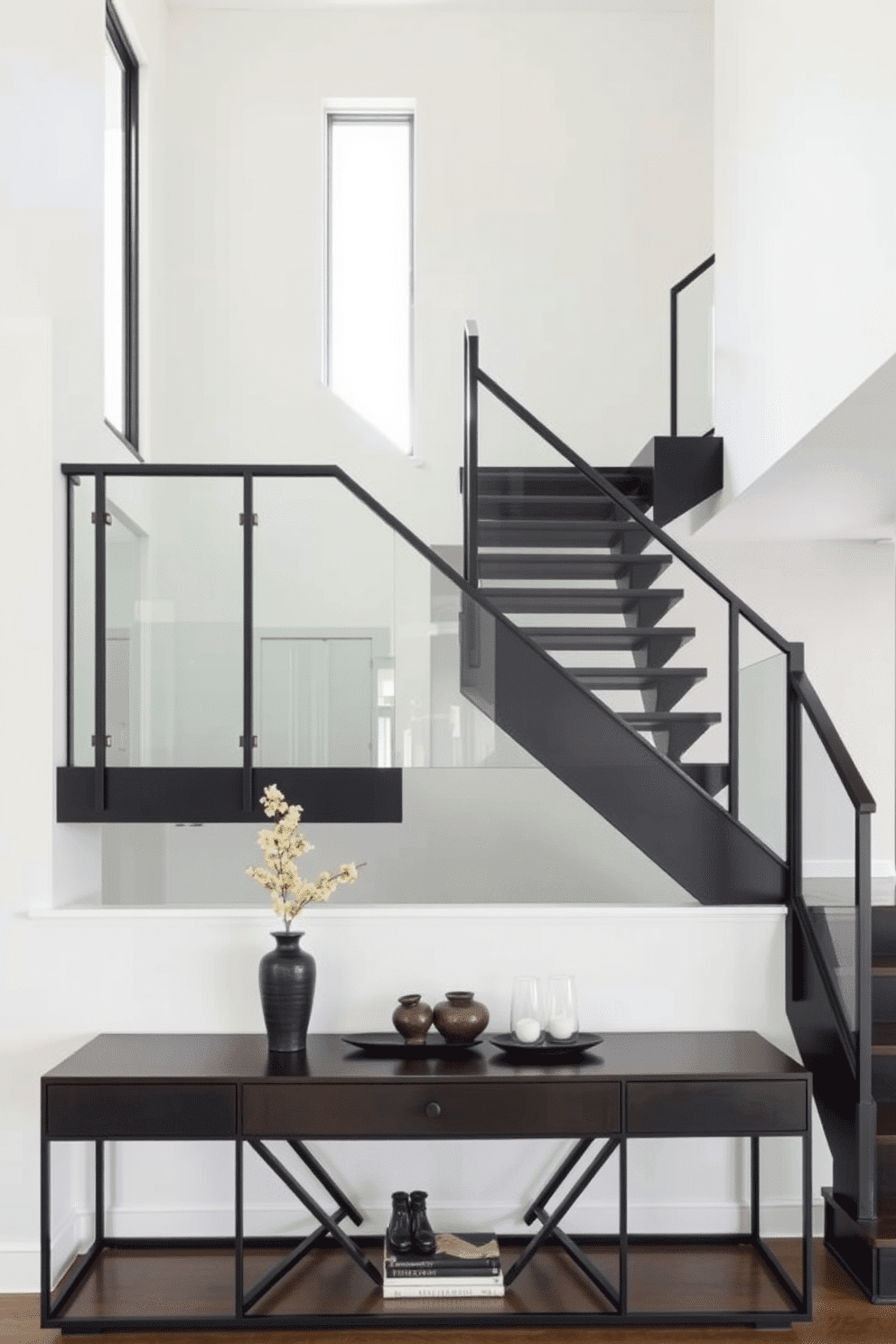 An open black staircase features sleek, minimalist lines that create a striking focal point in the space. The glass side panels add an element of transparency, allowing natural light to flow through while providing a modern touch. The staircase is set against a backdrop of light-colored walls, enhancing its dramatic presence. Below, a stylish console table with decorative accents complements the overall design, making the area feel inviting and sophisticated.