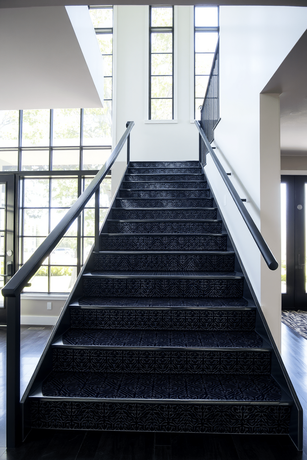 A textured black staircase features intricate patterned risers that add visual interest and depth. The sleek handrail complements the staircase's modern aesthetic, creating a striking focal point in the entryway. Surrounding the staircase, natural light floods in through large windows, highlighting the unique textures and patterns. The contrast between the black staircase and the bright walls enhances the overall elegance of the space.