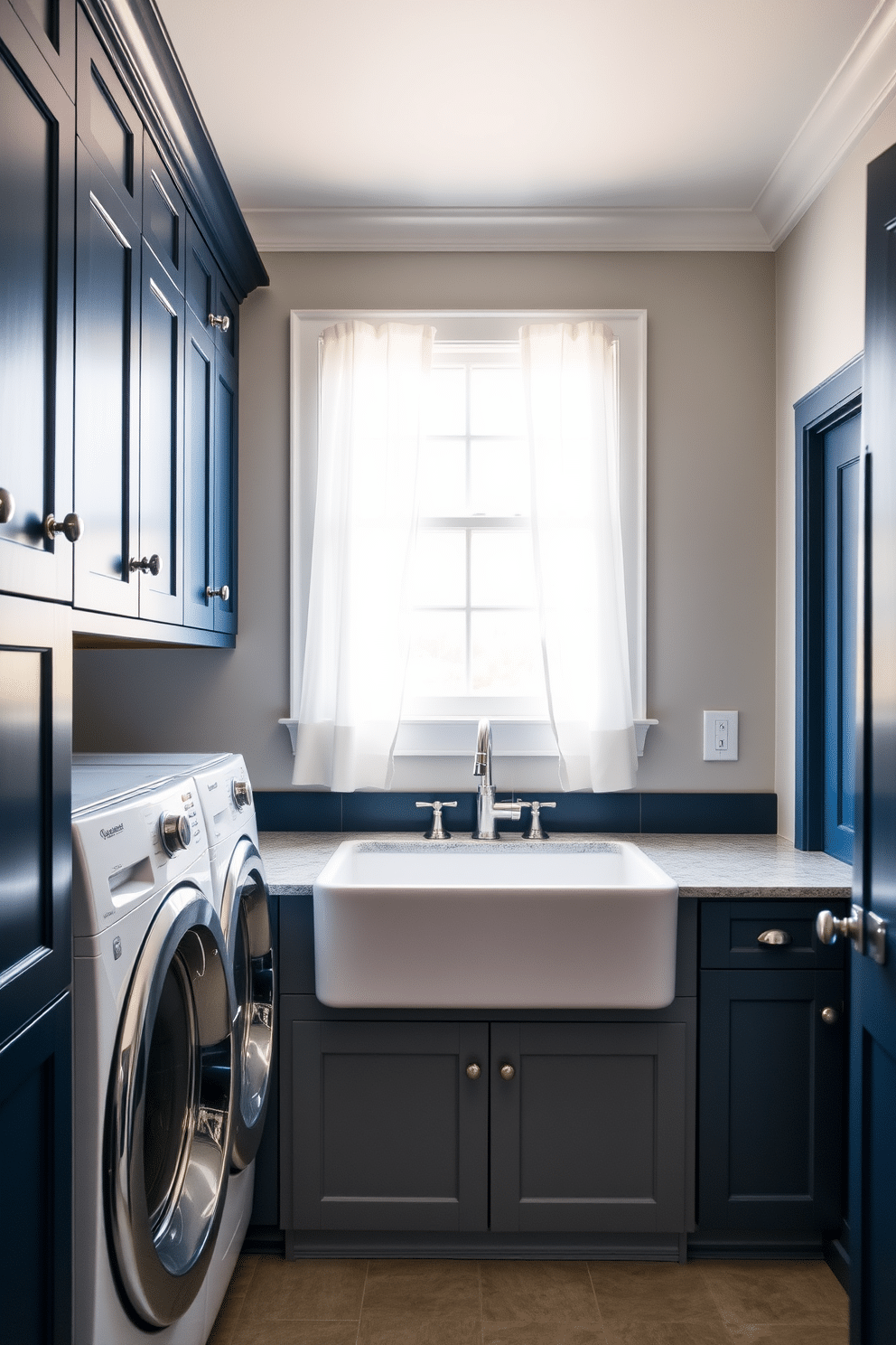 A sophisticated laundry room featuring a blue and gray color scheme. The walls are painted a soft gray, while the cabinets are a deep navy blue, providing a striking contrast. The laundry appliances are seamlessly integrated with the cabinetry, accented by brushed nickel hardware. A large farmhouse sink sits beneath a window, framed by sheer white curtains that allow natural light to fill the space.