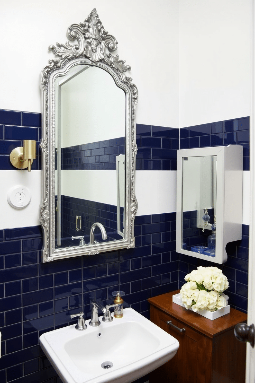 A chic powder room featuring navy blue tiles elegantly framing a large, ornate mirror. The walls are painted in a soft white, creating a striking contrast with the deep blue tiles, and a sleek pedestal sink complements the design. To the side, a small wooden cabinet provides storage while maintaining an airy feel. Decorative accents, such as a stylish soap dispenser and a fresh bouquet of white flowers, enhance the room's sophistication.