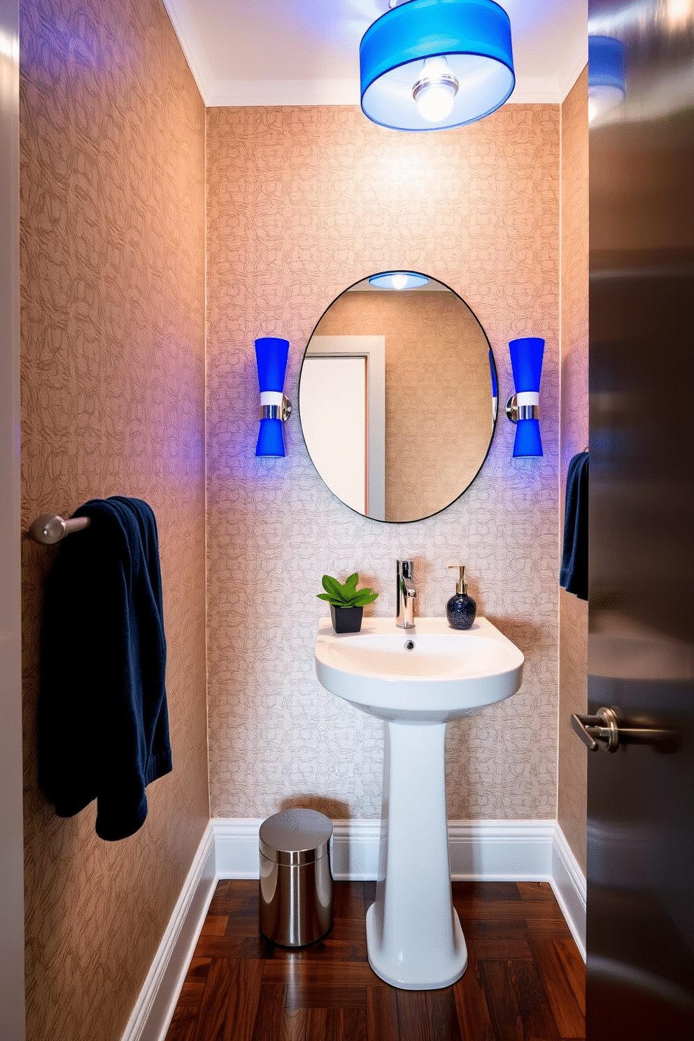 A chic powder room featuring unique blue light fixtures that cast a soft, inviting glow throughout the space. The walls are adorned with elegant wallpaper in a subtle pattern, complemented by a sleek white pedestal sink and a stylish round mirror above it. The floor is covered in rich, dark wood, creating a warm contrast with the blue accents. Decorative elements include a small potted plant on the sink and a plush, navy blue towel hanging on a polished chrome rack.