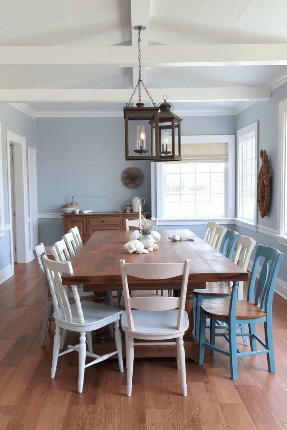 A cozy coastal dining room features a large reclaimed wood table surrounded by mismatched chairs, each painted in soft pastel hues. Above the table, rustic lanterns hang from a beam, casting a warm glow that enhances the room's inviting atmosphere. The walls are adorned with light blue shiplap, complemented by white trim and large windows that let in natural light. Nautical decor, such as seashells and driftwood accents, adds a charming touch to the overall design.