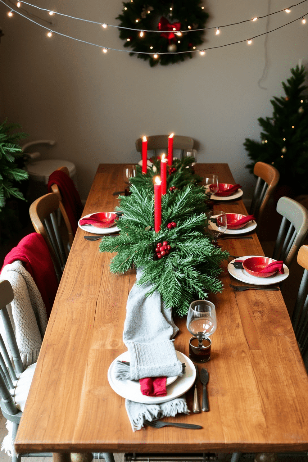 A rustic wooden table is adorned with lush evergreen centerpieces, creating a warm and inviting atmosphere. The table is surrounded by mismatched chairs, each draped with soft, textured throws to enhance the cozy holiday feel. Twinkling fairy lights are strung above, casting a gentle glow over the scene. Festive tableware featuring red and green accents complements the natural elements, adding a cheerful touch to the dining experience.