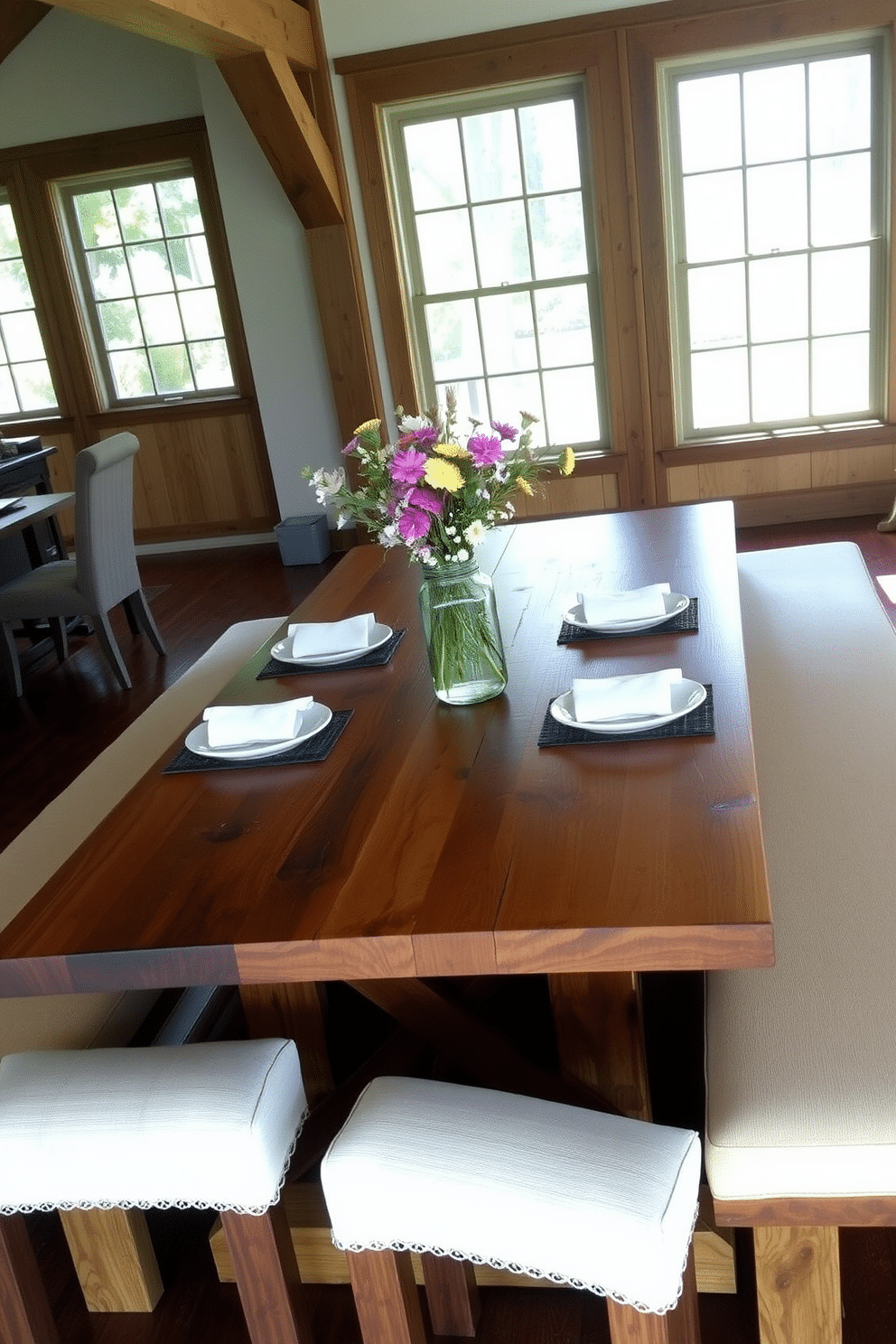 A rustic farmhouse dining table made of reclaimed wood, showcasing natural knots and a weathered finish. Flanking the table, there are long bench seats upholstered in soft, neutral fabric, creating a cozy and inviting atmosphere. The room features exposed wooden beams overhead and large windows that let in ample natural light. A centerpiece of wildflowers in a mason jar sits atop the table, complemented by vintage-style place settings.