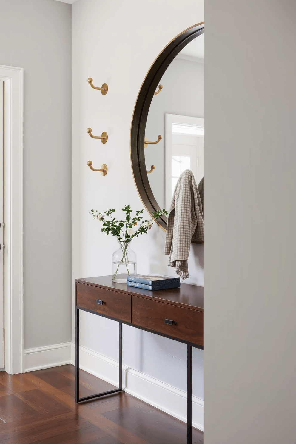 A stylish entryway featuring wall-mounted hooks made of brushed brass, providing a modern touch while ensuring easy organization. The walls are painted a soft gray, and a sleek console table with a dark wood finish sits beneath a large round mirror, reflecting natural light into the space.