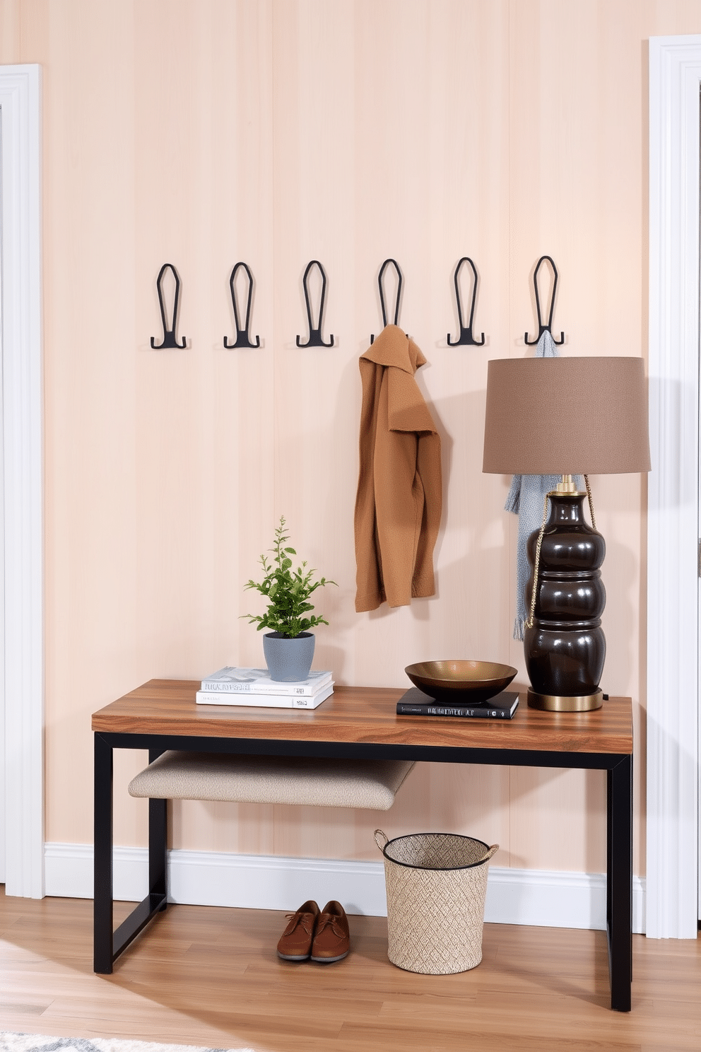 A modern entryway features a set of sleek, minimalist coat hooks mounted on a light wood wall, each designed with a unique geometric shape. Below the hooks, a plush bench upholstered in a neutral fabric provides a comfortable spot for putting on shoes, while a small potted plant adds a touch of greenery. The entryway table showcases a blend of contemporary and rustic elements, with a reclaimed wood top and black metal legs. Adorning the table are decorative items such as a stylish lamp, a stack of coffee table books, and a chic bowl for keys, creating an inviting and organized space.