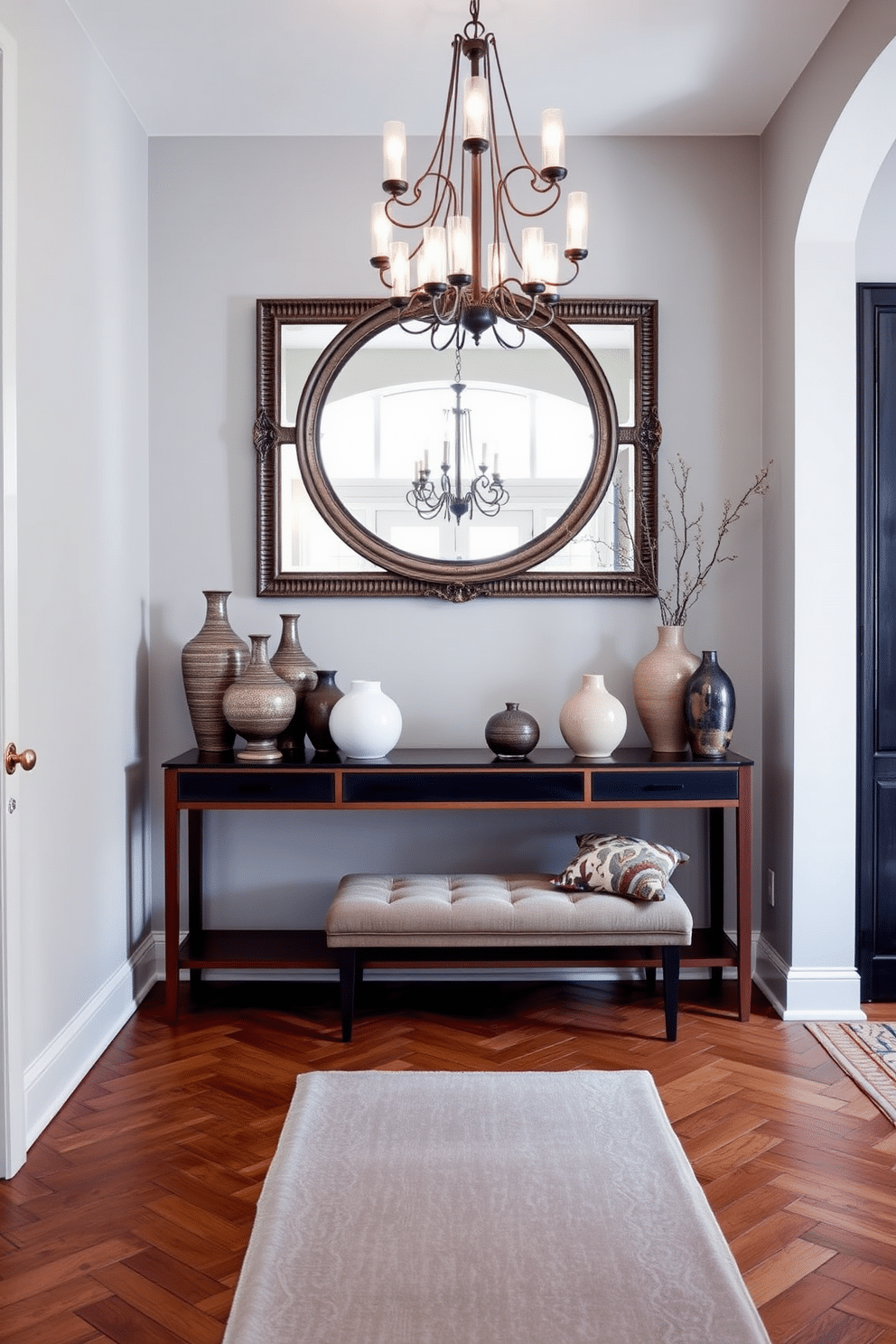 A stylish console table is positioned against a light gray wall, adorned with an array of decorative vases in varying heights and textures. The floor features a classic herringbone pattern in warm wood tones, complemented by a large, ornate mirror above the table that reflects natural light. The foyer is illuminated by a contemporary chandelier with soft, ambient lighting, creating an inviting atmosphere. A plush area rug in muted colors anchors the space, while a small bench with decorative cushions provides a functional yet stylish seating option.