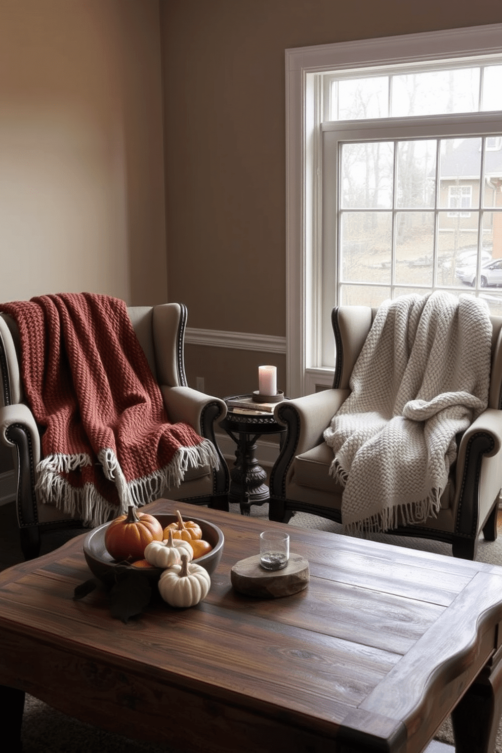 Textured blankets are casually draped over a pair of elegant armchairs in a cozy living room setting. The chairs are positioned near a large window that lets in soft, natural light, enhancing the warm tones of the blankets. A rustic wooden coffee table sits in front of the chairs, adorned with seasonal decor like small pumpkins and candles. The walls are painted in a warm beige, complementing the rich colors of the blankets and creating an inviting atmosphere.