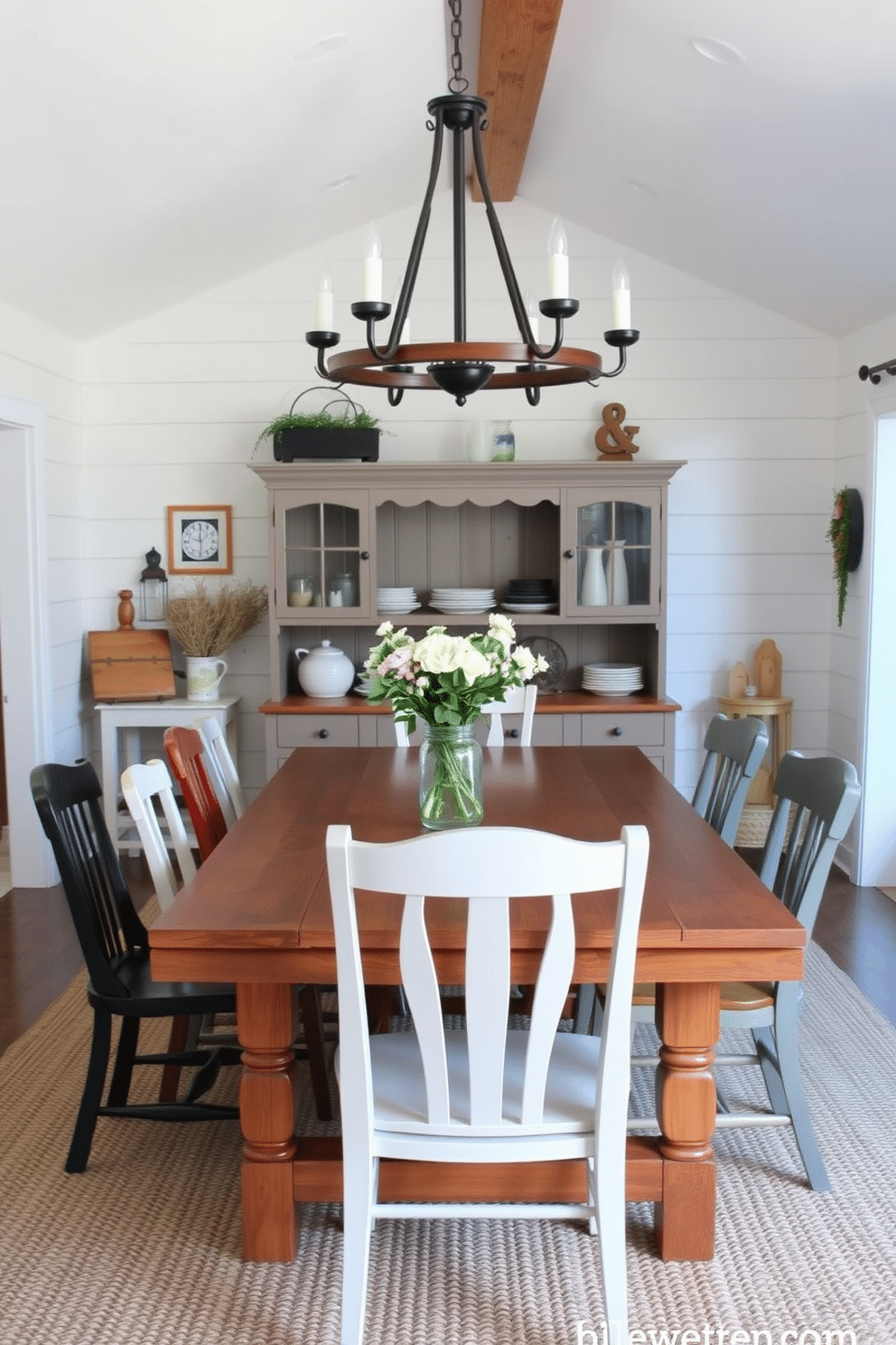 A charming farmhouse dining room features a large wooden table surrounded by mismatched chairs, creating a warm and inviting atmosphere. Above the table, a rustic chandelier made of wrought iron and wood casts a soft glow, enhancing the cozy ambiance of the space. The walls are adorned with shiplap paneling painted in a soft white, complemented by vintage-inspired decor and a large farmhouse-style hutch filled with dishware. A woven rug underfoot adds texture, while fresh flowers in a mason jar serve as a centerpiece, bringing a touch of nature indoors.