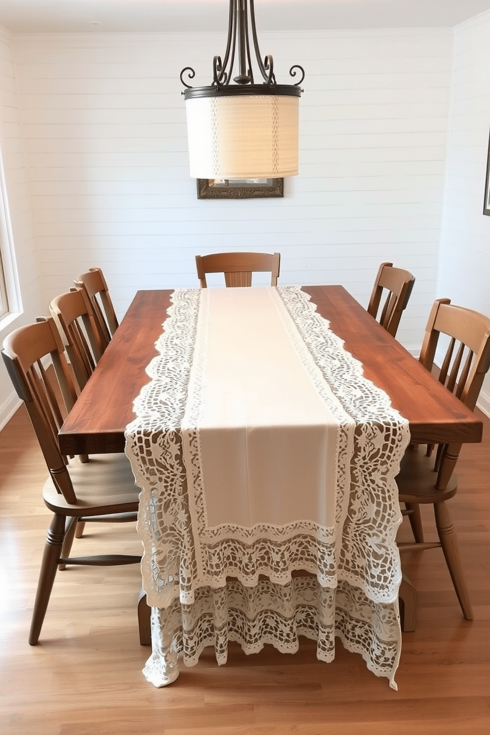 A vintage-inspired table runner drapes elegantly over a rustic farmhouse dining table, showcasing intricate lace details and a soft, muted color palette. Surrounding the table are mismatched wooden chairs, each with its own unique finish, creating a charming and inviting atmosphere. The walls of the dining room are adorned with shiplap, painted in a warm white, complementing the natural wood tones of the furniture. A large farmhouse-style chandelier hangs above the table, casting a warm glow over the space and enhancing the cozy, nostalgic feel.