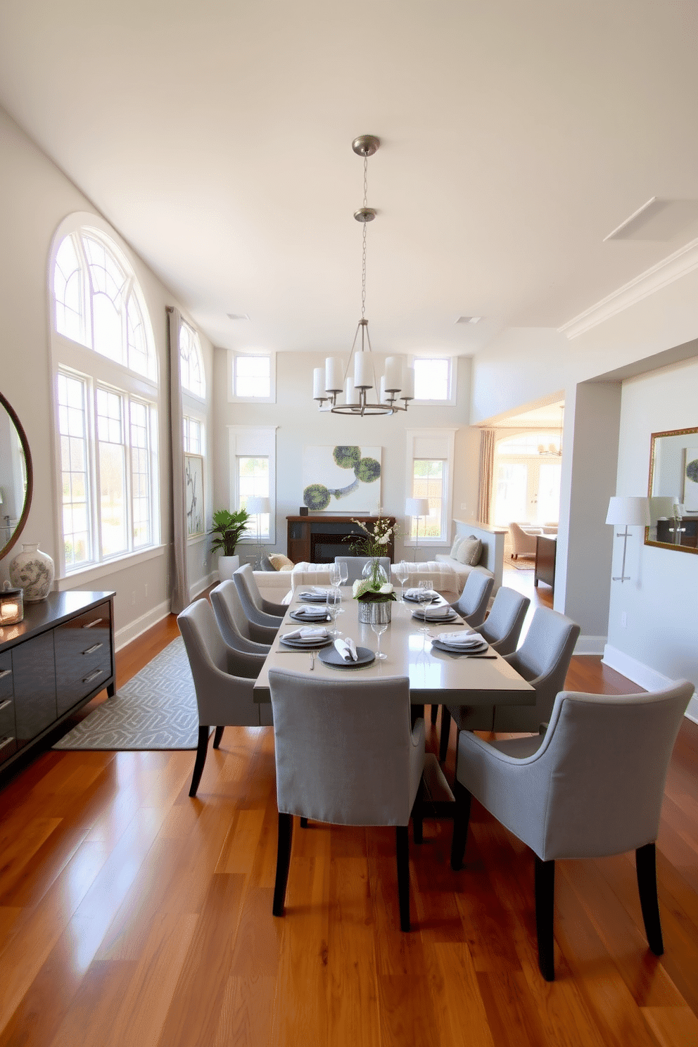 Open concept layout featuring a defined dining area with an elegant rectangular table set for eight, surrounded by upholstered chairs in a soft gray fabric. The space is illuminated by a modern chandelier overhead, while large windows allow natural light to flood in, highlighting the warm wood flooring. The dining area is separated from the living space by a stylish area rug that complements the color scheme. A sideboard against the wall displays decorative items and serves as additional storage, enhancing the overall sophistication of the room.