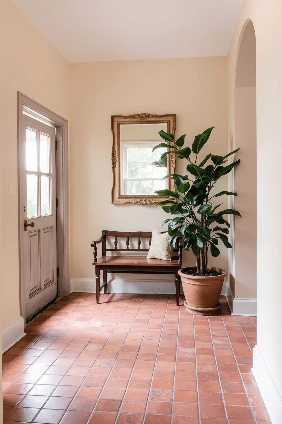 A welcoming foyer features rustic terracotta tiles that exude warmth and charm. The tiles are arranged in a herringbone pattern, complemented by a wooden bench and a large potted plant in the corner. Above the bench, a vintage mirror with a distressed frame reflects natural light coming through a nearby window. The walls are painted in a soft cream color, enhancing the earthy tones of the terracotta tiles.