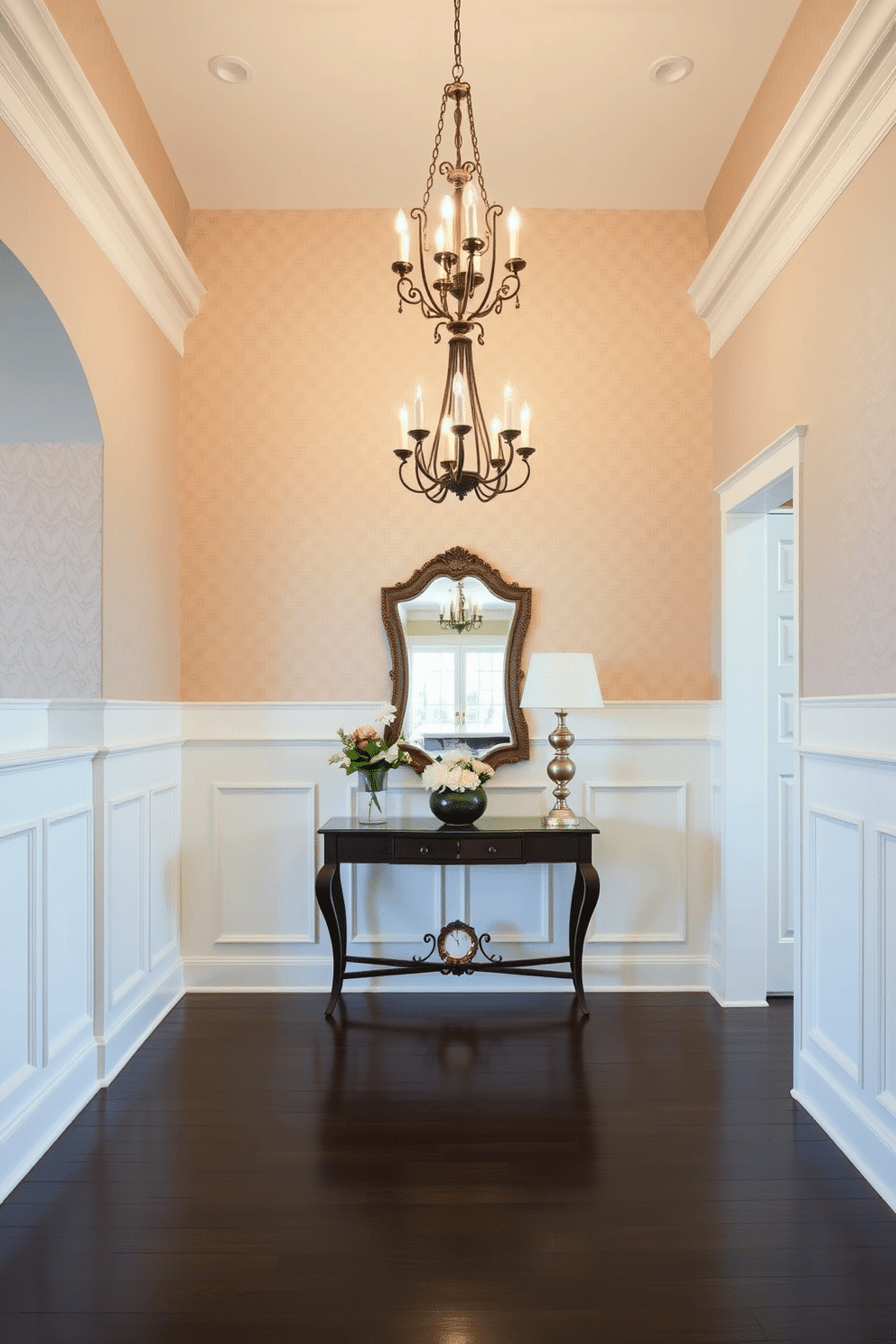 A grand foyer features elegant wainscoting that extends halfway up the walls, painted in a soft ivory color. The upper wall is adorned with a subtle, textured wallpaper that adds depth, while a classic chandelier hangs from the ceiling, illuminating the space. The foyer showcases a stylish console table against the wainscoted wall, topped with a decorative mirror and fresh flowers. Dark hardwood flooring complements the design, leading to a welcoming area that sets the tone for the rest of the home.