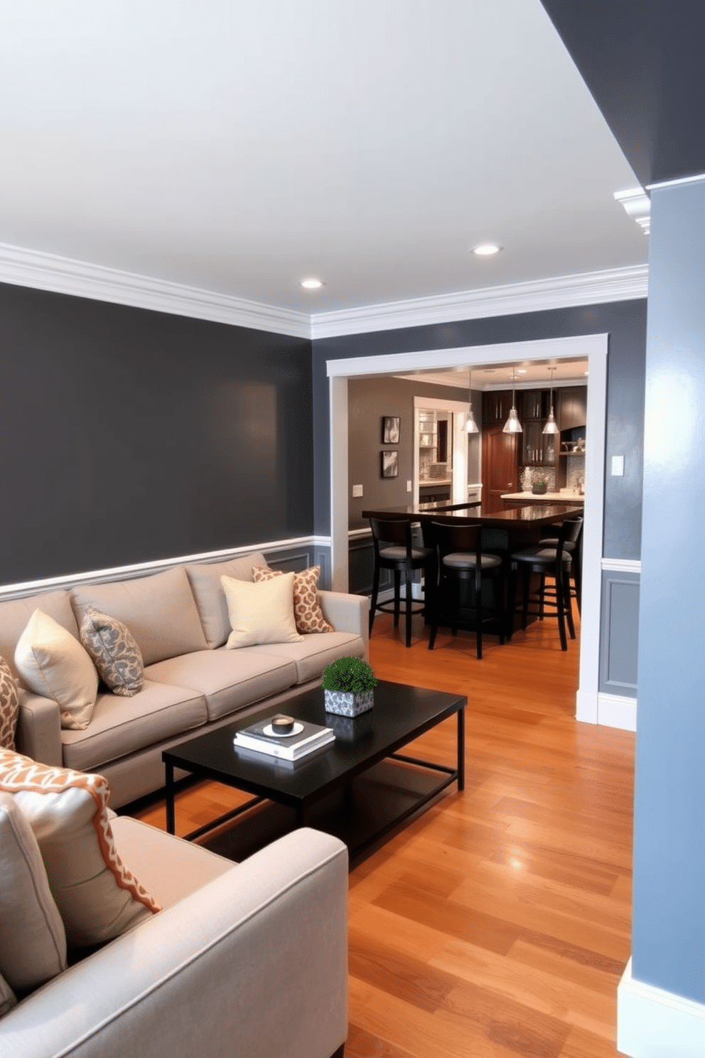 A stylish gray basement retreat featuring charcoal gray walls complemented by crisp white trim. The space includes a plush sectional sofa in light gray, accented with colorful throw pillows, and a sleek coffee table in the center. In one corner, a home bar area showcases a dark wood bar with high stools, illuminated by pendant lights. The flooring is a warm wood tone, creating a cozy contrast against the dark walls.