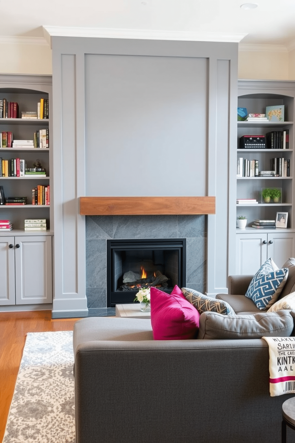 A modern gray fireplace with a sleek wood mantel serves as the focal point of the family room, surrounded by built-in shelves filled with books and decorative items. The space is adorned with a plush gray sectional sofa, complemented by vibrant accent pillows and a soft area rug that adds warmth to the hardwood floor.