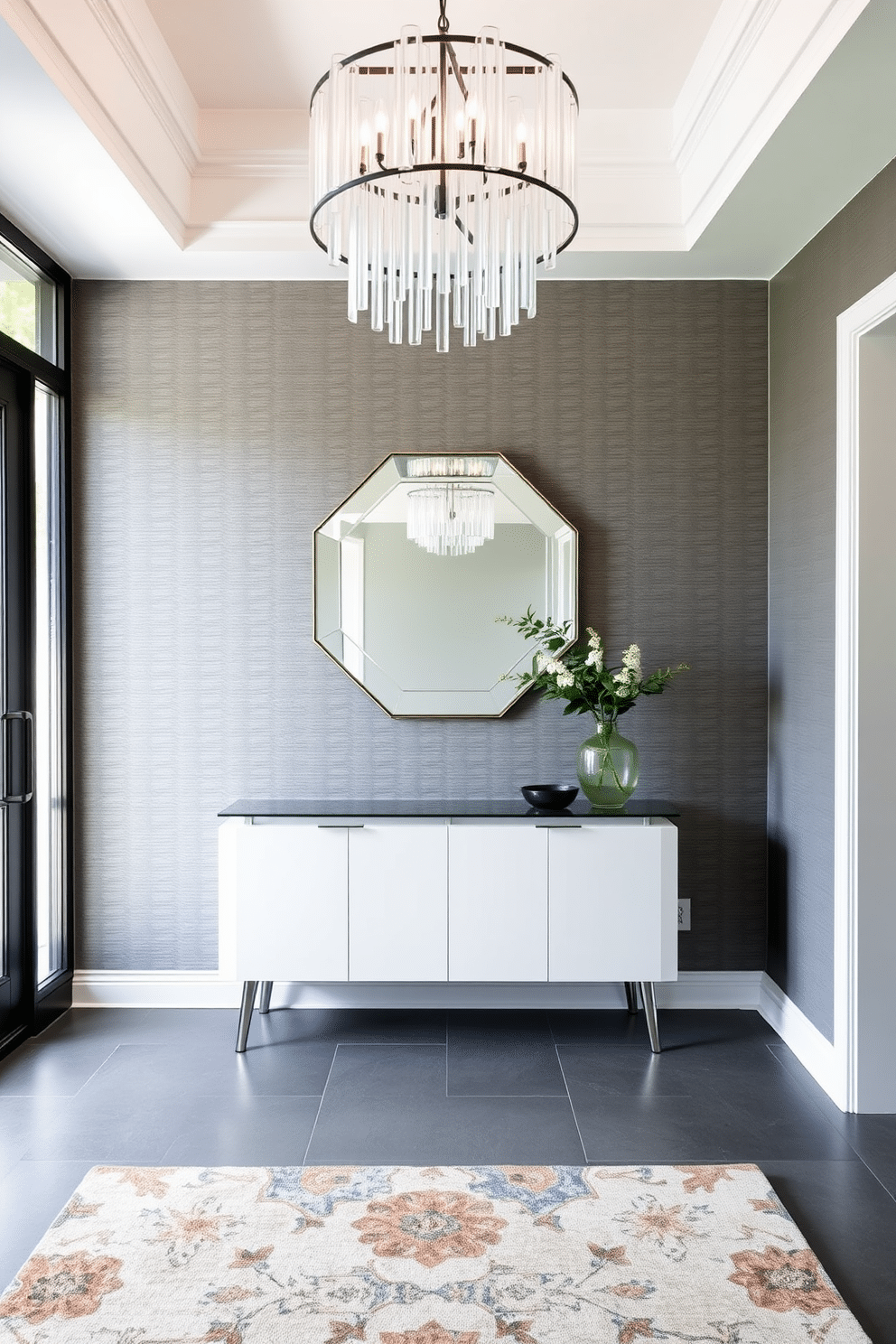 A sleek gray foyer featuring a large glass chandelier that cascades light across the space. The walls are adorned with a textured gray wallpaper, complemented by a minimalist console table with a glass top and elegant metal legs. A statement mirror with a geometric design hangs above the console, reflecting the natural light from the nearby window. The floor is finished with large-format gray tiles, and a stylish area rug adds warmth and texture to the entrance.