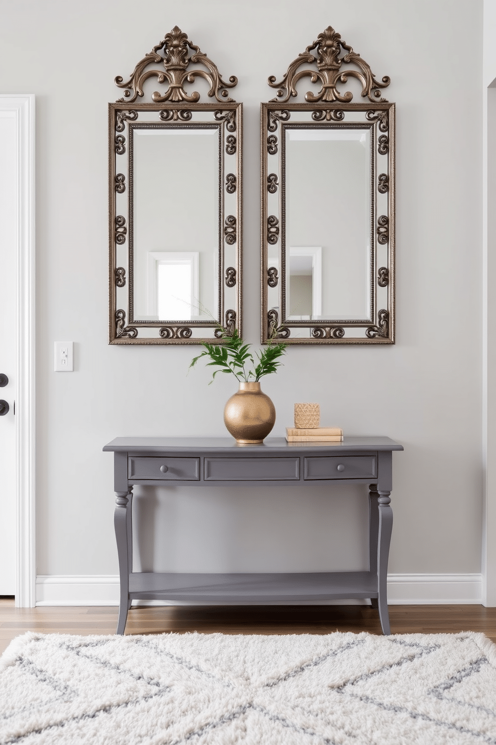 A chic gray console table is positioned against a light gray wall, reflecting the soft natural light from a nearby window. Above the table, a pair of elegant mirrors with ornate frames create a stunning focal point in the foyer. The floor is adorned with a plush, neutral-toned area rug that adds warmth to the space. Flanking the console table are stylish decorative accents, including a sculptural vase and a small potted plant, enhancing the overall aesthetic.