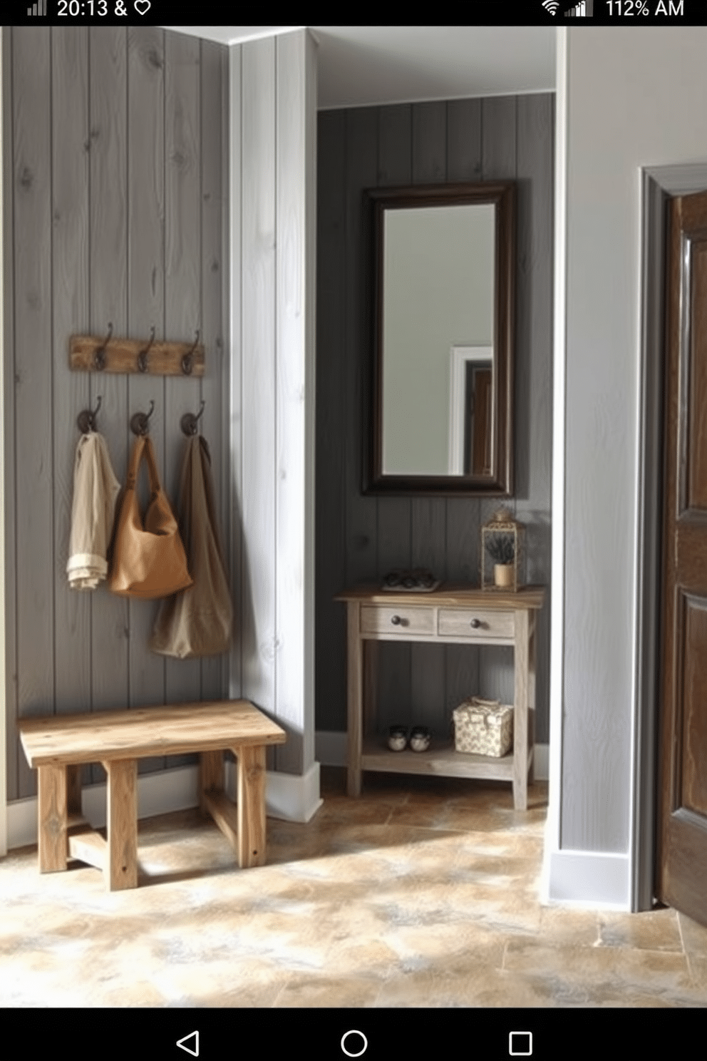 A cozy foyer featuring rustic gray wood paneling that adds warmth and character to the space. The floor is adorned with a natural stone tile, complementing the wood and creating an inviting atmosphere. To the left, a bench made of reclaimed wood offers a practical seating area, while a collection of decorative hooks on the wall provides storage for coats and bags. A large, framed mirror hangs above a small console table, reflecting light and enhancing the sense of space.