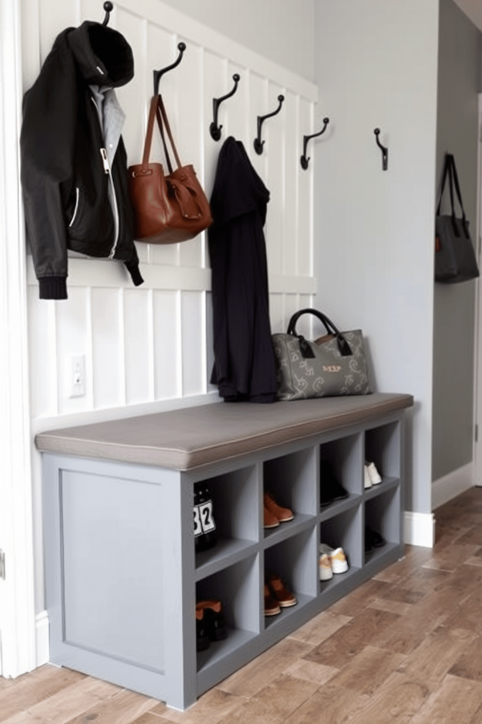 A functional gray bench with shoe storage is positioned against the wall in a stylish mudroom. The bench features a cushioned seat and a series of open cubbies below for easy access to shoes, while the walls are adorned with hooks for coats and bags.