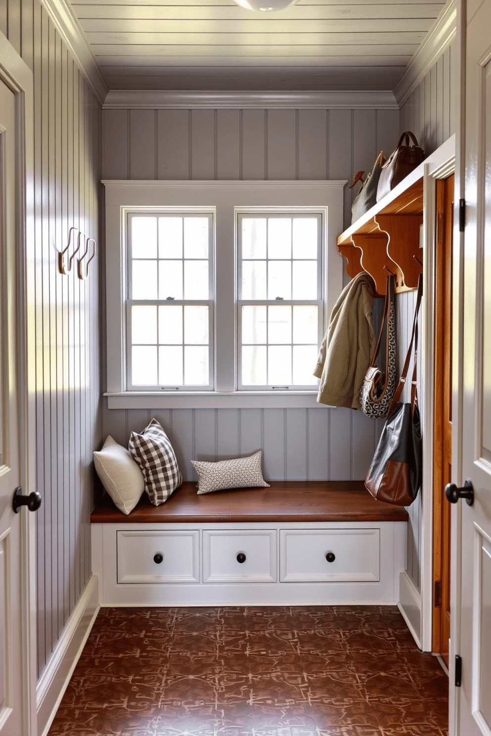 A cozy mudroom features gray painted beadboard paneling that adds a rustic charm. The space includes a built-in bench with soft cushions, surrounded by hooks for hanging coats and bags. Natural light floods in through a large window, illuminating the warm wood accents throughout. The floor is adorned with durable, patterned tiles that complement the gray beadboard beautifully.