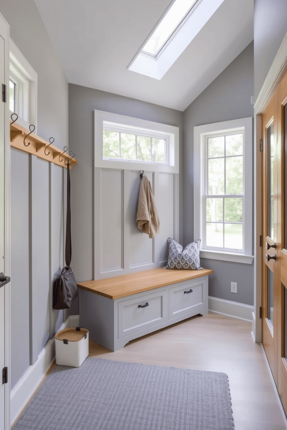 A stylish mudroom featuring a harmonious blend of gray and natural wood elements. The walls are painted in a soft gray hue, complemented by a warm wood bench with built-in storage underneath. To the left, a series of wooden hooks are mounted for hanging coats and bags, while a gray area rug adds texture to the floor. Large windows allow natural light to flood the space, enhancing the inviting atmosphere of this functional entryway.