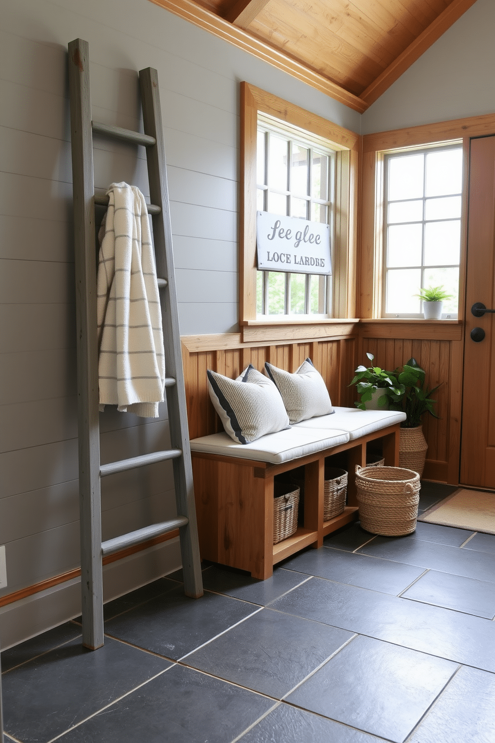 A rustic gray ladder leans against the wall, serving as a stylish towel storage solution in the mudroom. The space features warm wood accents, a durable slate floor, and a cozy bench with soft cushions, creating a welcoming atmosphere. The walls are adorned with shiplap in a soft gray hue, enhancing the rustic charm of the room. Natural light floods in through a large window, illuminating the carefully curated decor, including potted plants and woven baskets for added texture.