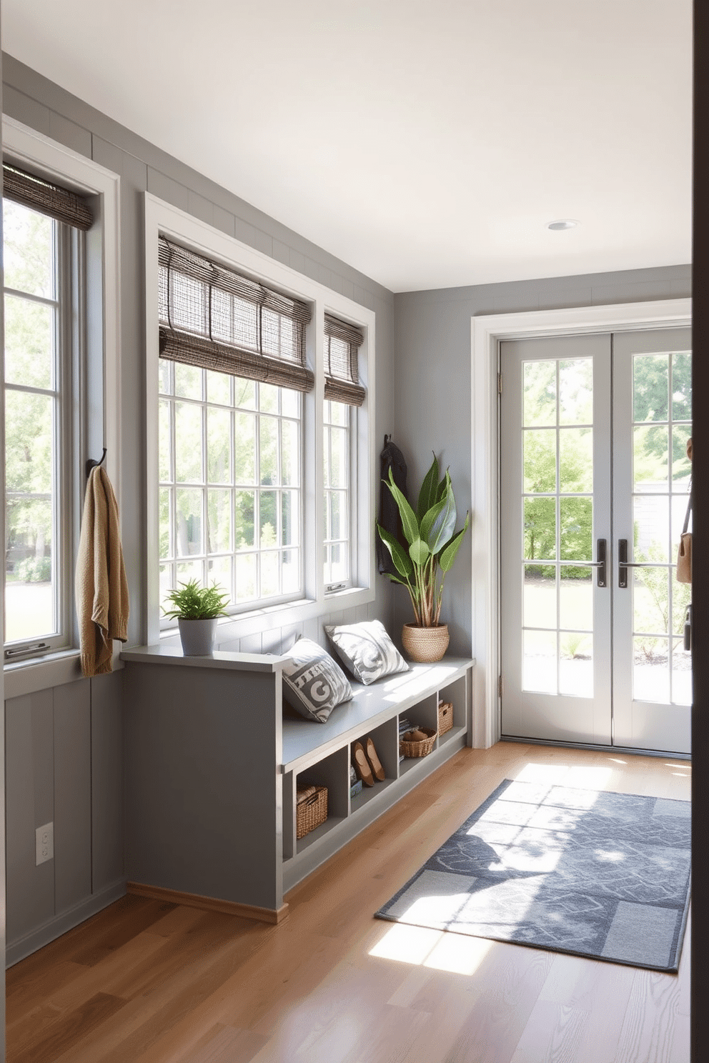 A bright gray mudroom featuring large windows that allow natural light to flood the space. The walls are painted in a soft gray, complemented by a built-in bench with storage underneath and hooks for coats above. The flooring is a durable light wood, providing warmth to the overall design. A stylish area rug adds a pop of color, while potted plants bring a touch of nature indoors.