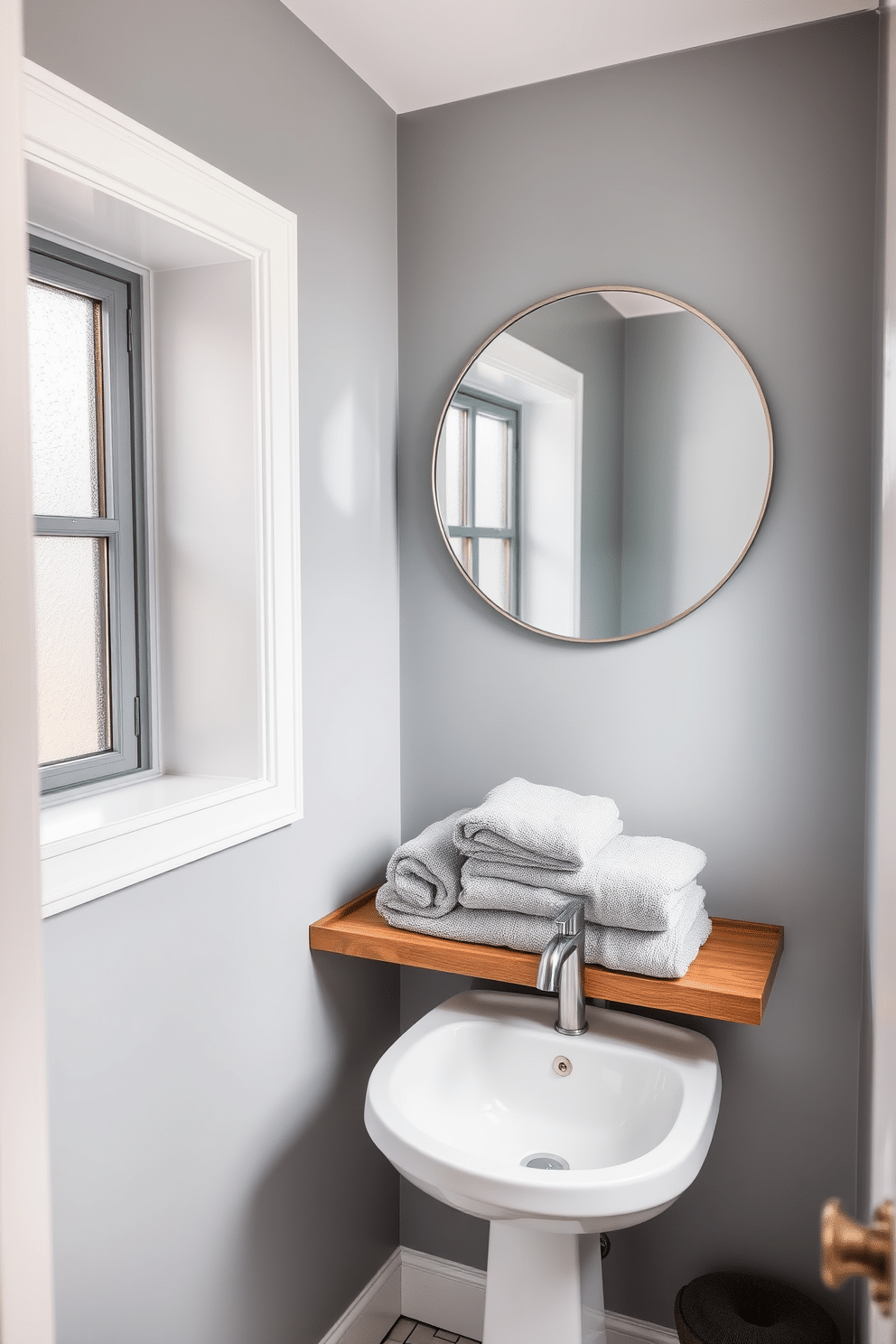 A serene powder room featuring soft gray walls that create a calming atmosphere. Plush gray towels are neatly arranged on a sleek wooden shelf, enhancing the spa-like ambiance. The space includes a stylish pedestal sink with a modern faucet, complemented by a round mirror framed in brushed nickel. Natural light filters in through a frosted window, illuminating the subtle textures of the decor.