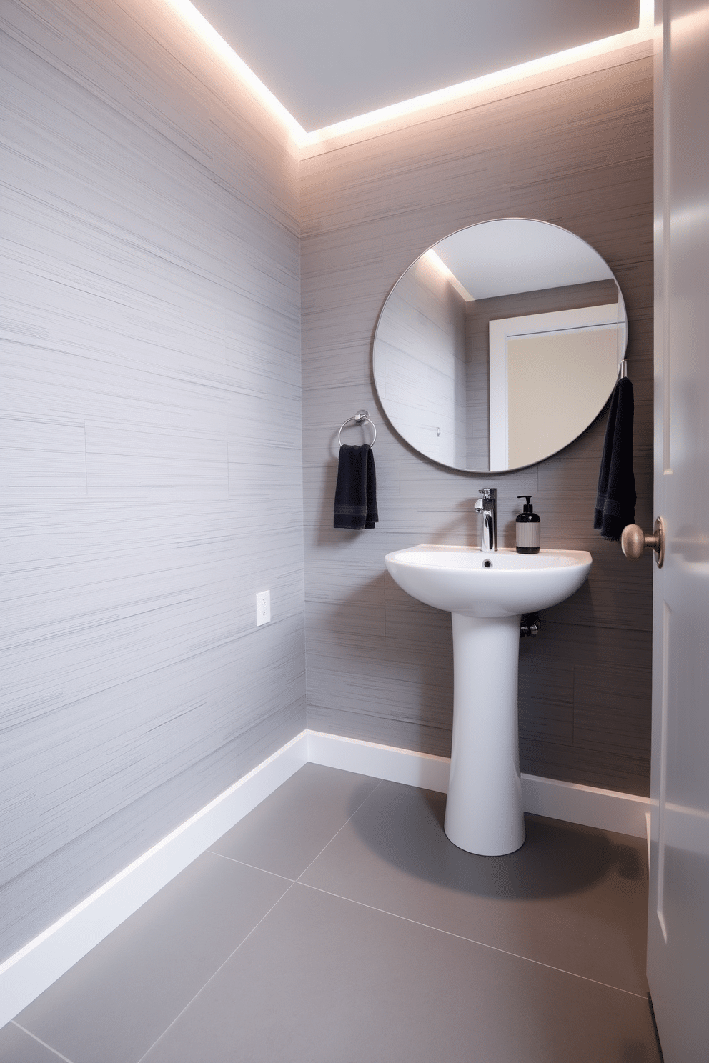 A chic powder room features sleek monochromatic gray tile flooring that creates a seamless and modern aesthetic. The walls are adorned with a subtle textured wallpaper in varying shades of gray, complementing the flooring while adding depth to the space. A minimalist pedestal sink with a polished chrome faucet stands against the wall, paired with a large round mirror that reflects the elegant lighting above. Accents of dark gray in the accessories, such as a stylish soap dispenser and a plush hand towel, enhance the sophisticated ambiance of the room.