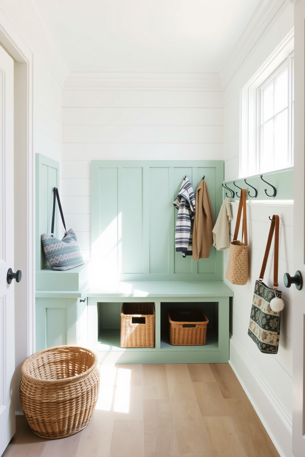 A seafoam green bench with built-in storage is the focal point of this mudroom, providing both functionality and style. The walls are adorned with shiplap in a soft white, creating a bright and airy atmosphere that complements the bench's color. To the right of the bench, a series of hooks are mounted for hanging coats and bags, while a woven basket sits beneath for additional organization. Natural light floods the space through a large window, enhancing the inviting feel of this well-designed mudroom.