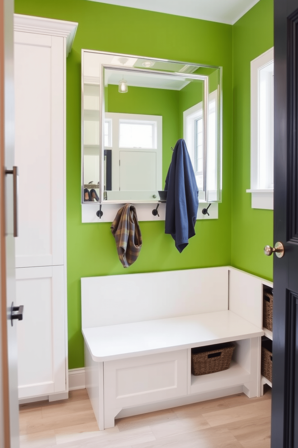 A bold green accent wall serves as the focal point of the mudroom, creating a vibrant contrast against the light-colored cabinetry. A large, stylish mirror is mounted above a sleek bench, providing both functionality and a touch of elegance. The space features built-in storage solutions with hooks for coats and baskets for shoes, all designed to keep the area organized and clutter-free. Natural light floods in through a nearby window, enhancing the welcoming atmosphere of this functional yet chic mudroom.