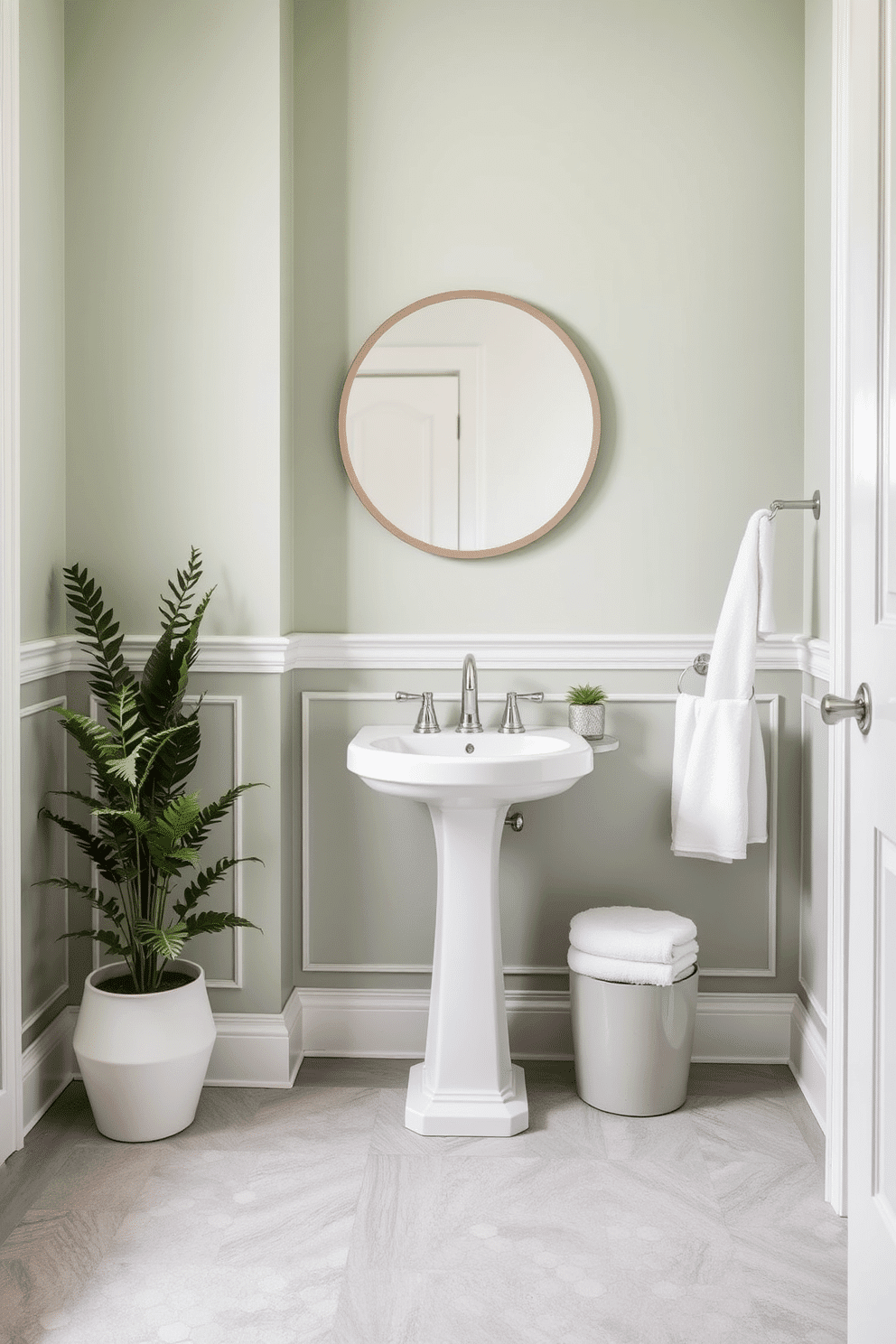 Soft sage walls with white trim create a serene and inviting atmosphere. A sleek white pedestal sink is paired with a round mirror framed in brushed nickel, reflecting light beautifully. The floor features elegant hexagonal tiles in a light gray hue, adding a touch of modern sophistication. Accents of greenery, such as a potted fern, bring life to the space, while a plush white towel is neatly hung on a stylish towel rack.