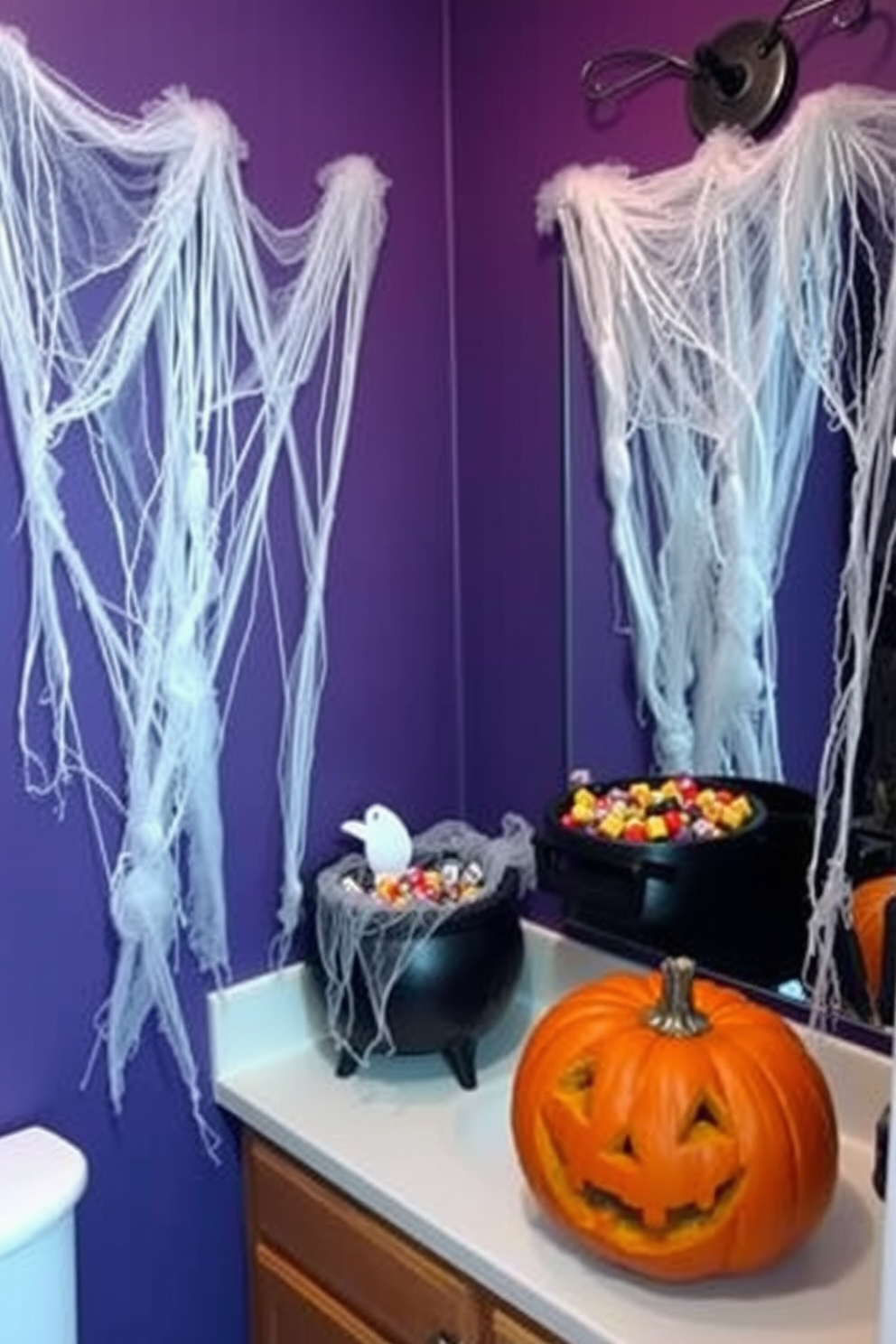 A spooky Halloween bathroom setting with faux cobwebs draped over the mirrors creating an eerie atmosphere. The walls are painted in deep purple, and a black cauldron filled with candy sits on the countertop next to a carved pumpkin.