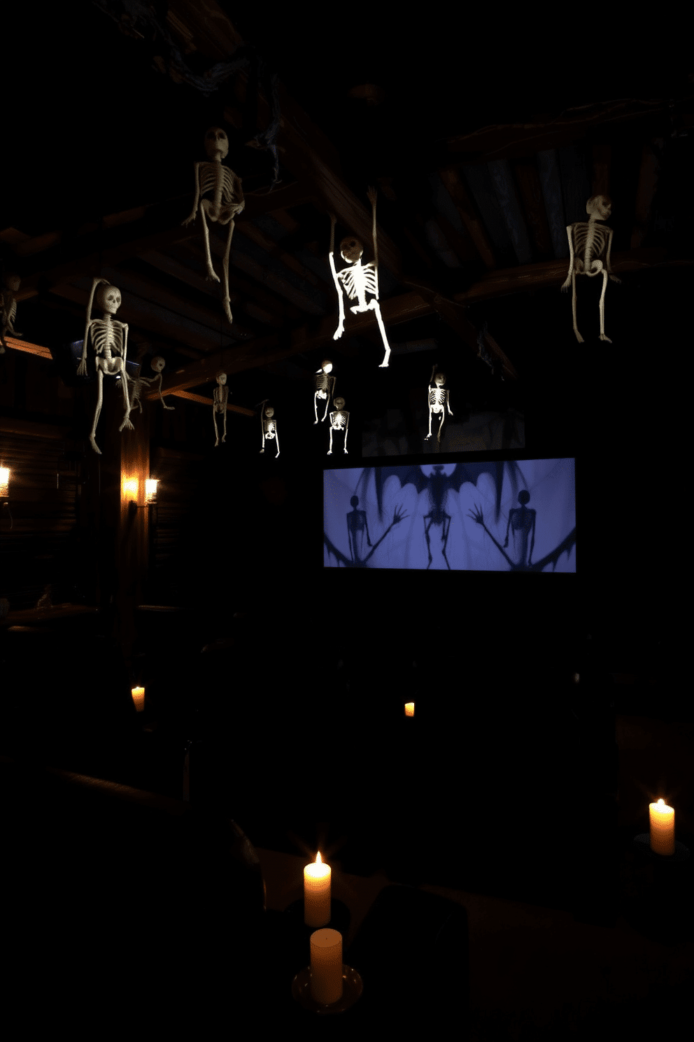 A cozy home theater setting for Halloween. Skeletons dangle from rustic ceiling beams, casting playful shadows on the walls. The seating area features plush black recliners arranged in a semi-circle. Dim lighting enhances the spooky atmosphere, with flickering candles placed strategically around the room.