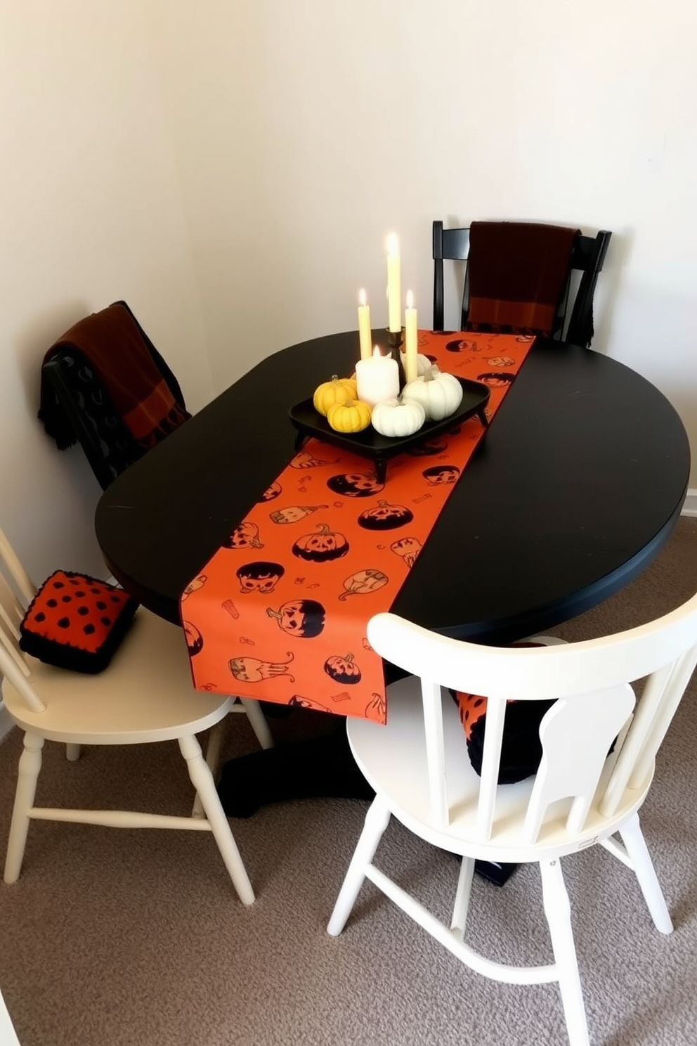 A cozy dining nook adorned with a black table topped with an orange table runner featuring Halloween motifs. Surrounding the table are four mismatched chairs, each draped with a small black and orange throw blanket for added warmth. On the table, a centerpiece of miniature pumpkins and flickering candles creates an inviting atmosphere. The walls are painted a soft cream color, enhancing the autumnal decor while maintaining a spacious feel in the small area.