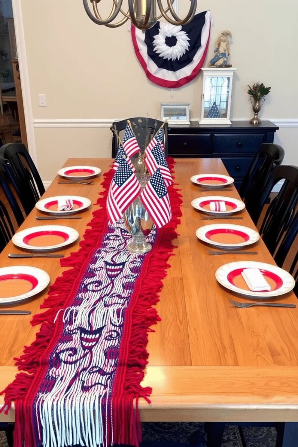 A festive dining room setting featuring a long wooden table adorned with patriotic table runners that have fringe accents. The table is set with red, white, and blue dinnerware, and small American flags are placed in decorative vases along the center.