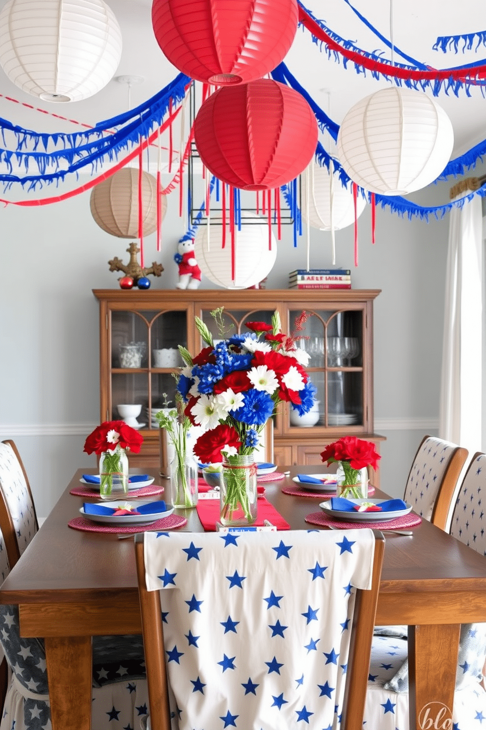 A vibrant dining room setting inspired by firecrackers. The table is adorned with colorful centerpieces featuring red white and blue flowers arranged in rustic mason jars. Festive decorations hang from the ceiling in the form of streamers and paper lanterns. The dining chairs are draped with star patterned fabric to enhance the Independence Day theme.