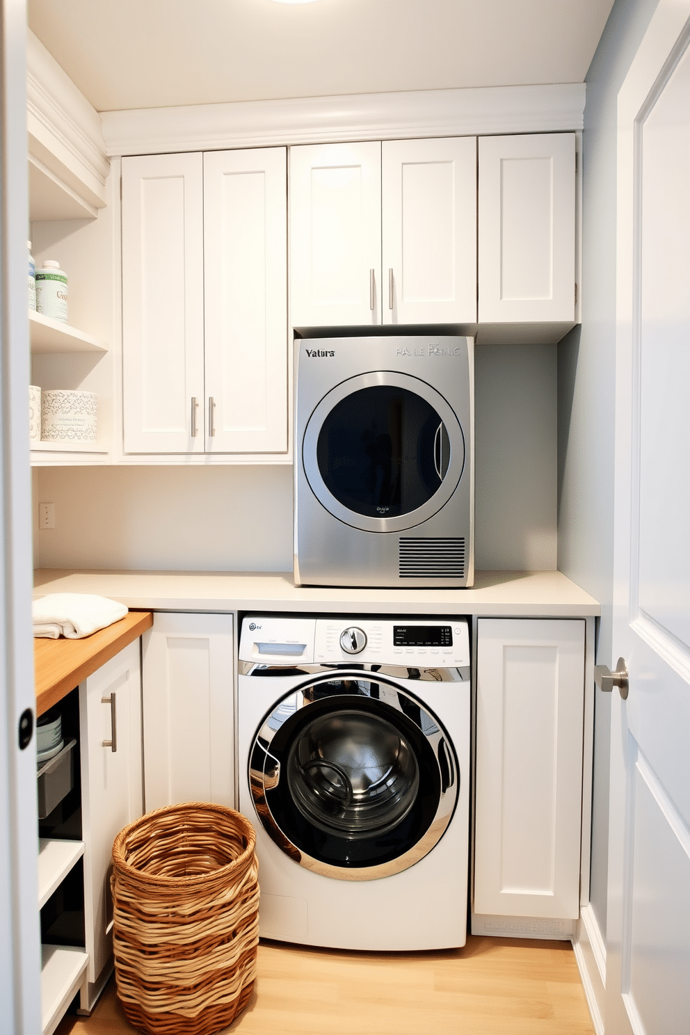 A modern kitchen laundry room featuring a stacked washer and dryer seamlessly integrated under a sleek countertop. The cabinetry is a crisp white with brushed nickel hardware, and the walls are painted in a soft gray, creating a clean and airy atmosphere. To the left of the appliances, a small folding station is designed with a wooden countertop, complemented by stylish open shelving above for easy access to laundry essentials. A decorative basket for linens sits on the floor, adding a touch of warmth and organization to the space.