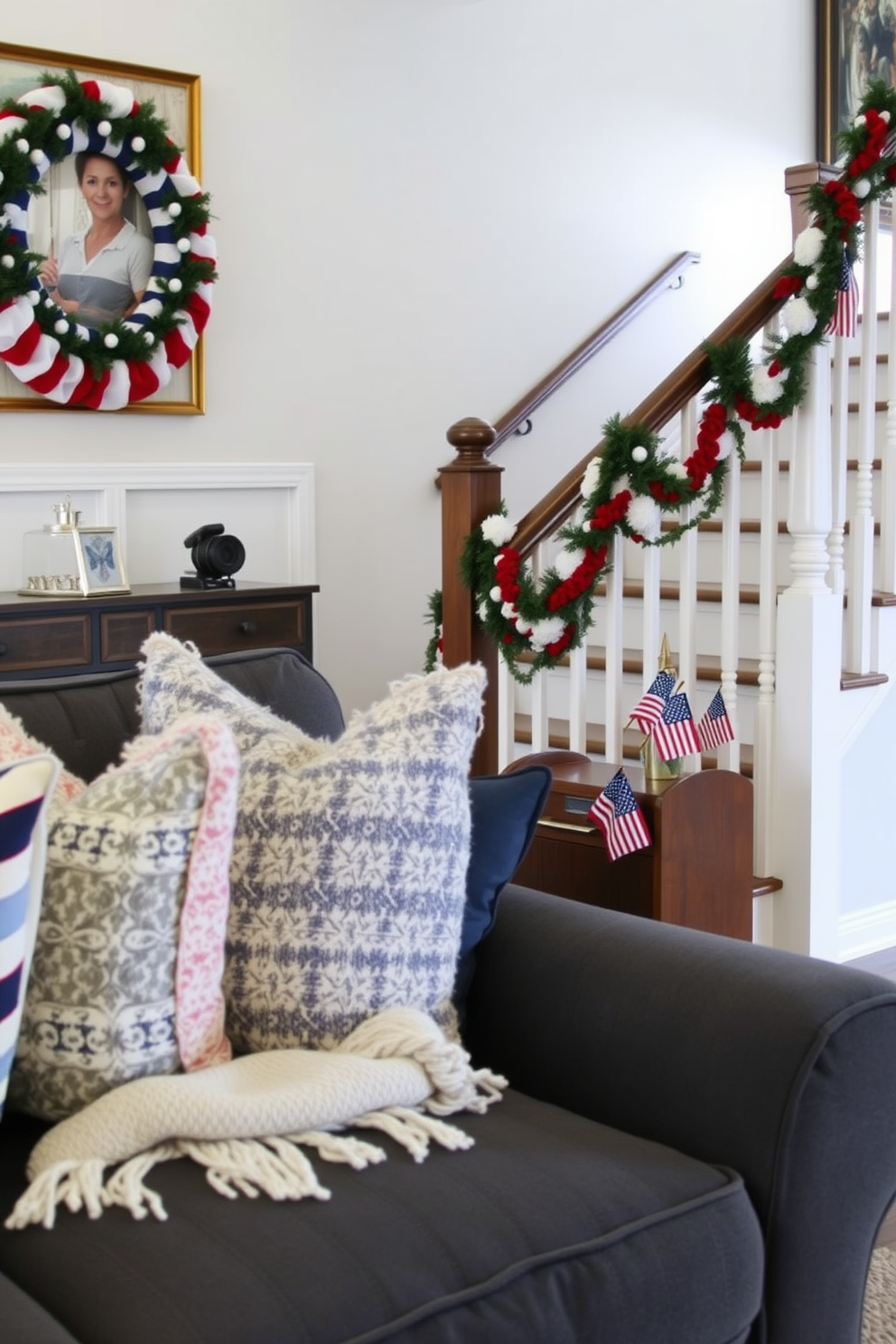 A cozy living room featuring layered textiles that create warmth and style. Plush throw pillows in various patterns and textures adorn a deep gray sofa, while a soft knitted blanket is draped over the armrest. The staircase is elegantly decorated for Labor Day with red, white, and blue accents. Festive garlands intertwine along the banister, and small American flags are placed strategically on the steps for a patriotic touch.