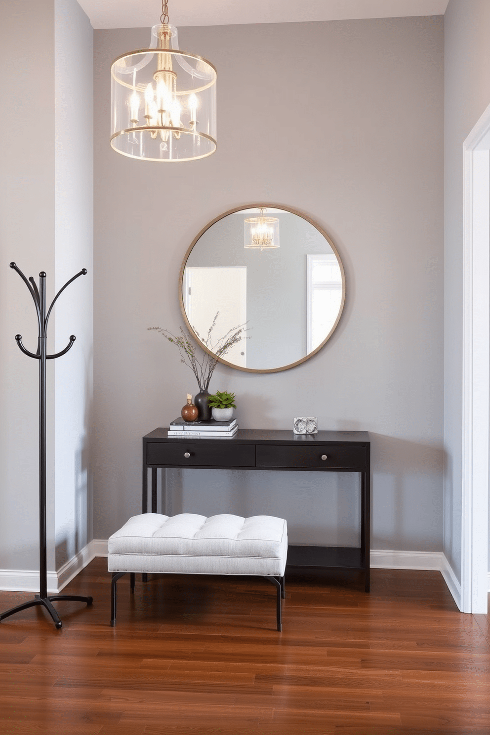 A chic coat rack stands against a backdrop of light gray walls, crafted from sleek metal with a minimalist design that complements the space. Below the rack, a stylish bench upholstered in a soft fabric offers a comfortable spot to sit while putting on or taking off shoes. The large entryway features an elegant chandelier that illuminates the space, casting a warm glow over the polished hardwood floor. A large round mirror hangs above a console table, adorned with decorative items and a small potted plant, creating an inviting atmosphere.