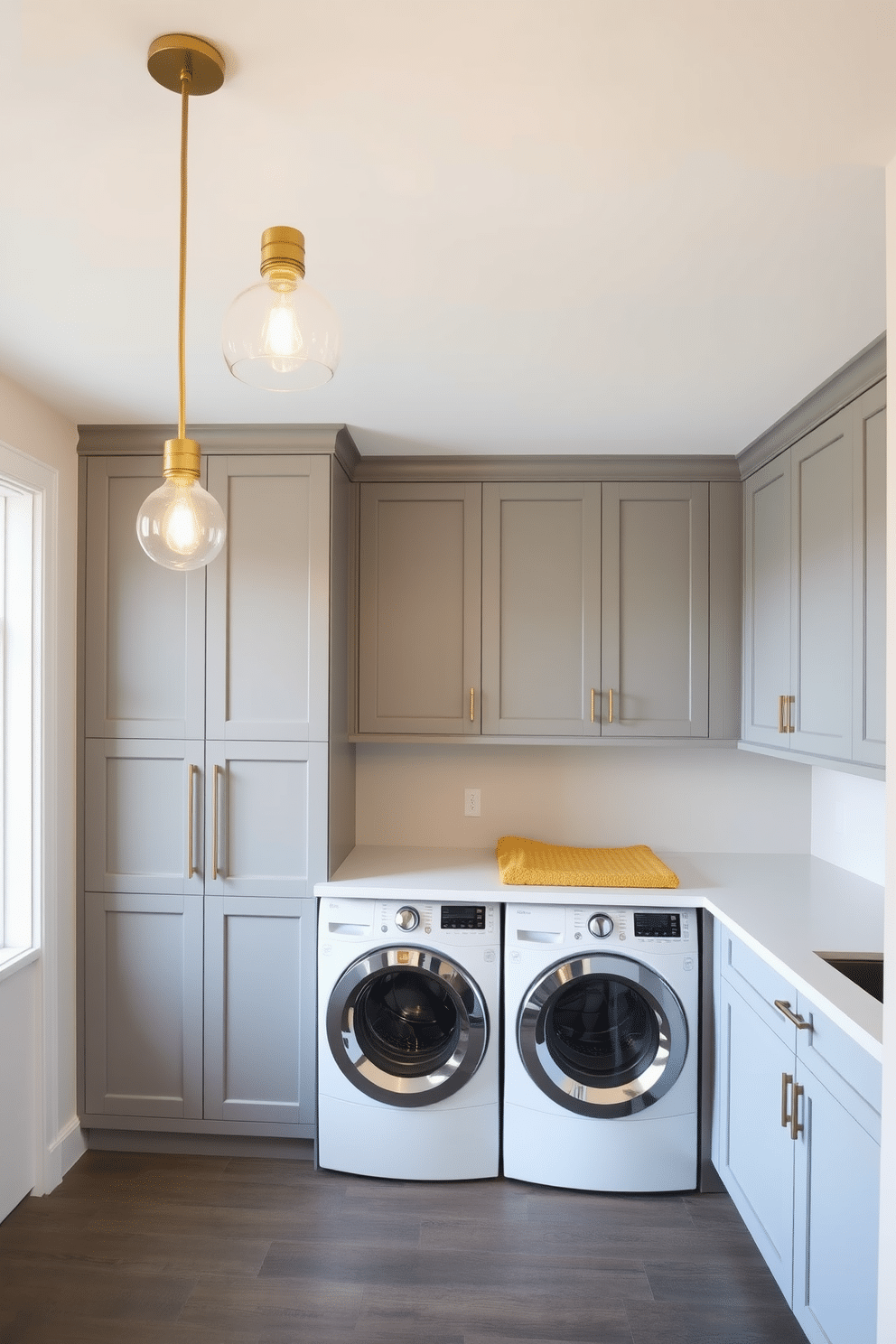 Chic pendant lighting hangs gracefully from the ceiling, casting a warm glow over the space. The fixtures feature a modern design with brass accents that complement the overall decor. The laundry room is designed with a sleek, functional layout, featuring built-in cabinetry in a soft gray finish. A large countertop for folding clothes is positioned beside the washer and dryer, creating an organized and stylish environment.