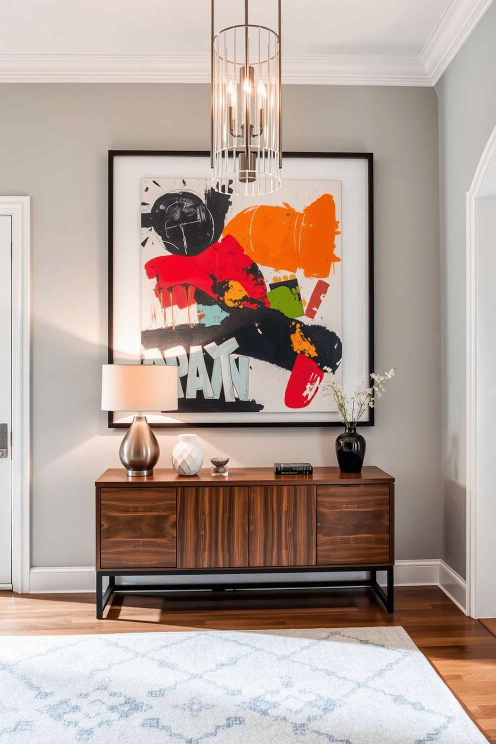 A stunning living room entryway features oversized artwork that commands attention and adds dramatic flair. The artwork, a vibrant abstract piece, is framed in a sleek black frame and is mounted on a light gray wall, creating a striking contrast. Beneath the artwork, a minimalist console table in a rich walnut finish showcases decorative objects and a stylish lamp. The entryway is illuminated by a modern chandelier, casting a warm glow that enhances the inviting atmosphere of the space.