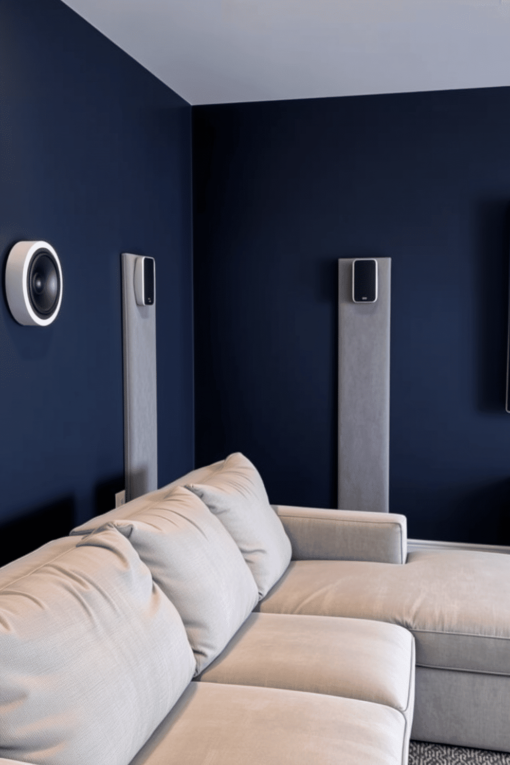 A contemporary media game room featuring wall-mounted speakers strategically placed for optimal surround sound. The walls are painted in a deep navy blue, complemented by a plush sectional sofa in a light gray fabric, perfect for lounging during movie nights or gaming sessions.