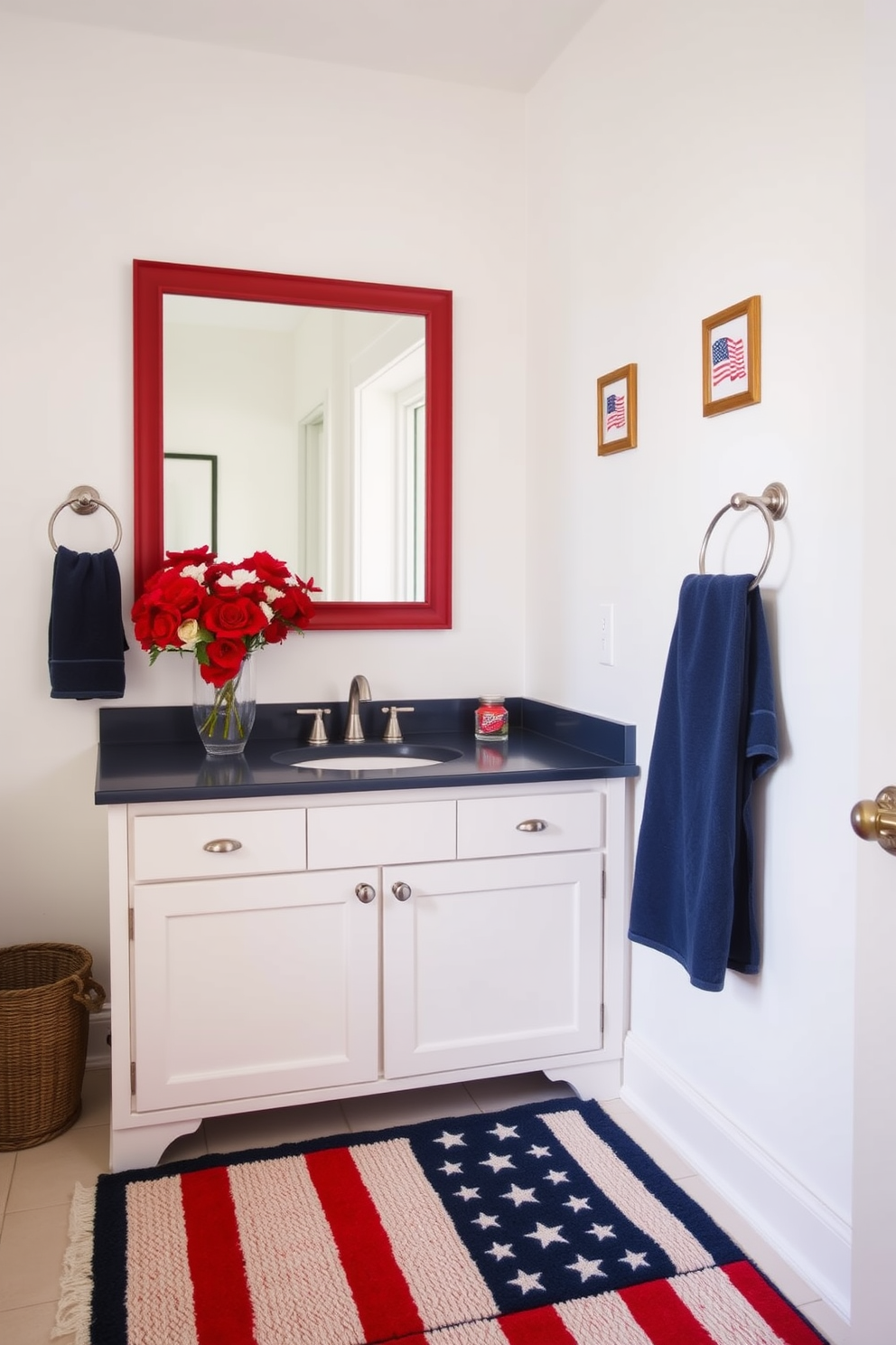 A patriotic bathroom setting. A red, white, and blue bath rug lies in front of a white vanity with a navy blue countertop. Above the vanity, a large rectangular mirror with a red frame reflects the light from a nearby window. The walls are painted a crisp white, adorned with small framed pictures of American flags. On the countertop, a vase filled with red and white flowers adds a festive touch. A navy blue towel hangs neatly from a silver towel rack beside the vanity.